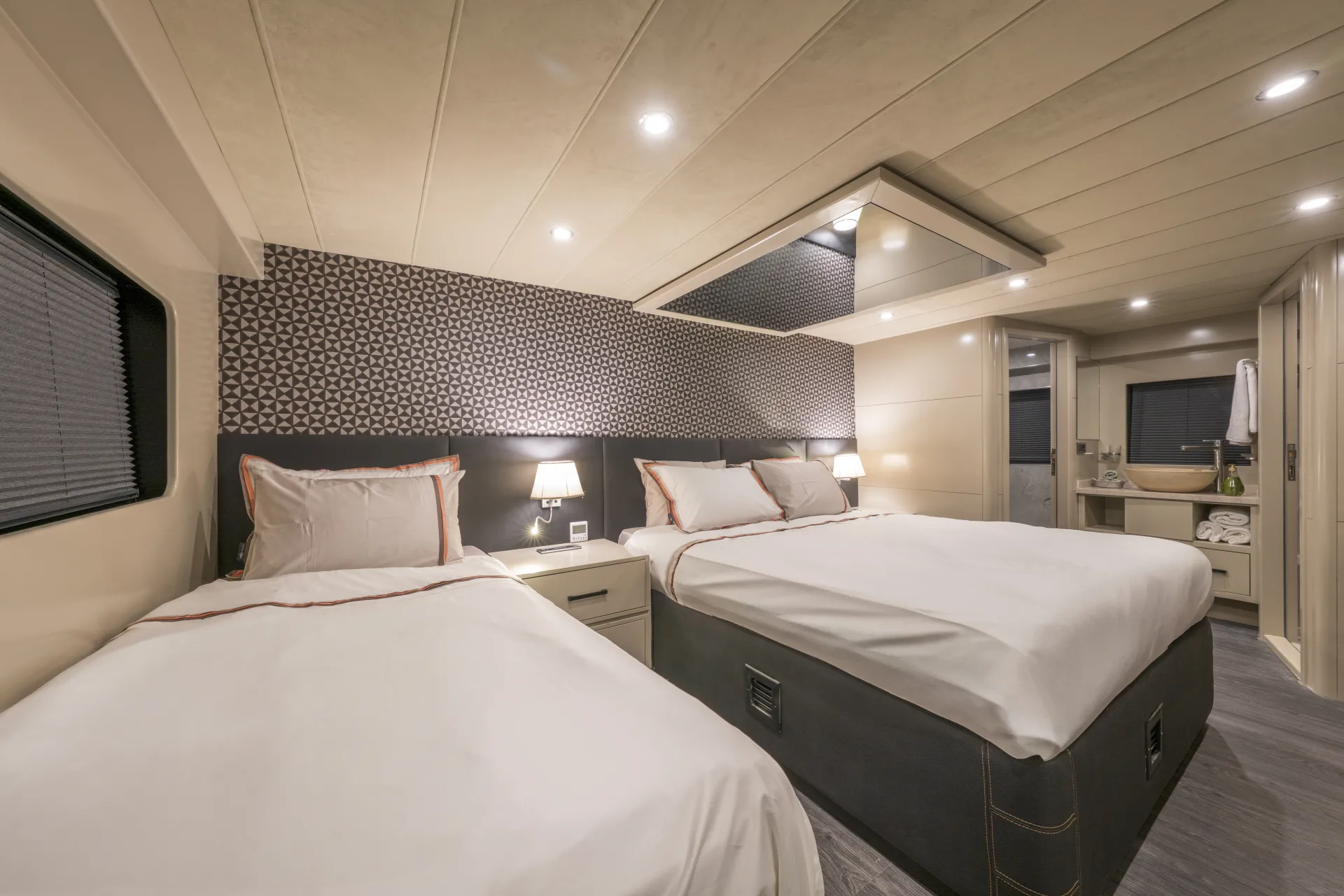 JUNIOR SIMAY Modern bedroom with two beds, white bedding, geometric patterned accent wall, bedside lamps, and an en suite bathroom visible in the background. The room has warm lighting and a clean, contemporary design.