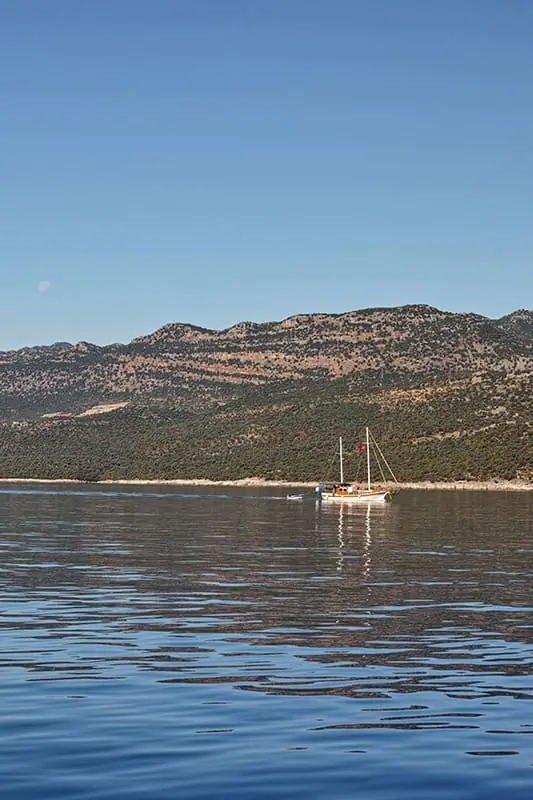 A small sailboat with a red flag floats on calm, reflective water near a forested shoreline with hills under a clear blue sky.