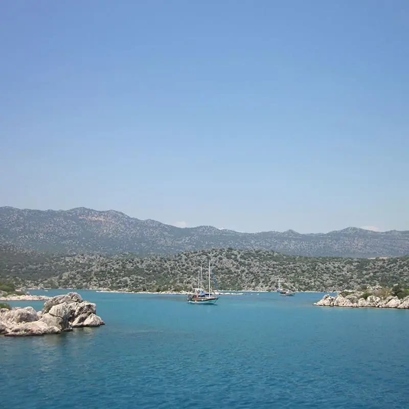 A sailboat floats on calm blue water surrounded by rocky shores and distant green hills under a clear blue sky.