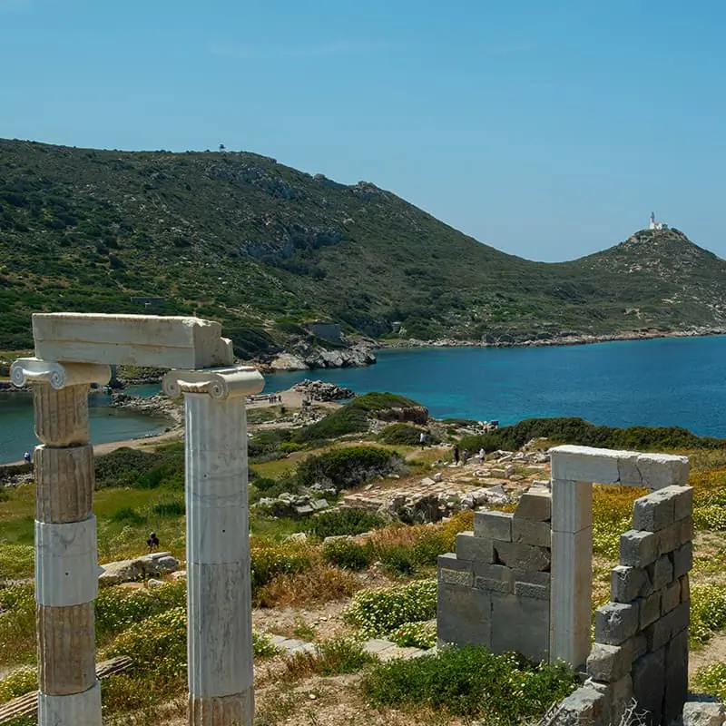 Ancient stone columns and arch stand near blue water, with green hills and a lighthouse in the distance under a clear sky.
