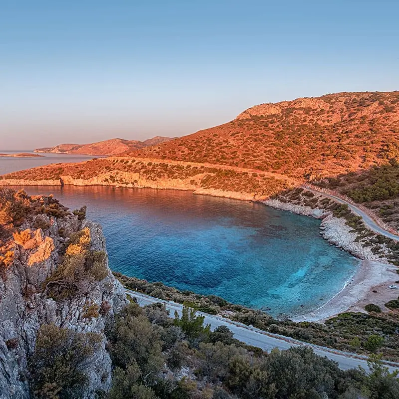 Rocky coastline with a small, clear blue bay, surrounded by hills under a clear sky at sunset. A winding road runs along the hillside above the shoreline. Sparse vegetation covers the area.