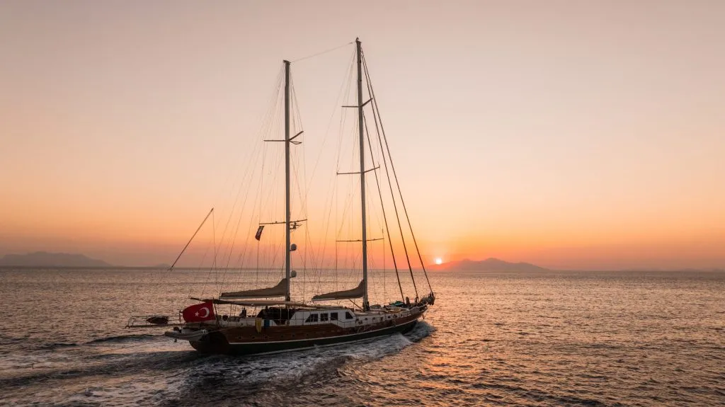 A large sailboat with multiple masts sails on calm water at sunset, with mountains in the background and the sun low on the horizon. A Turkish flag is visible at the back of the boat.