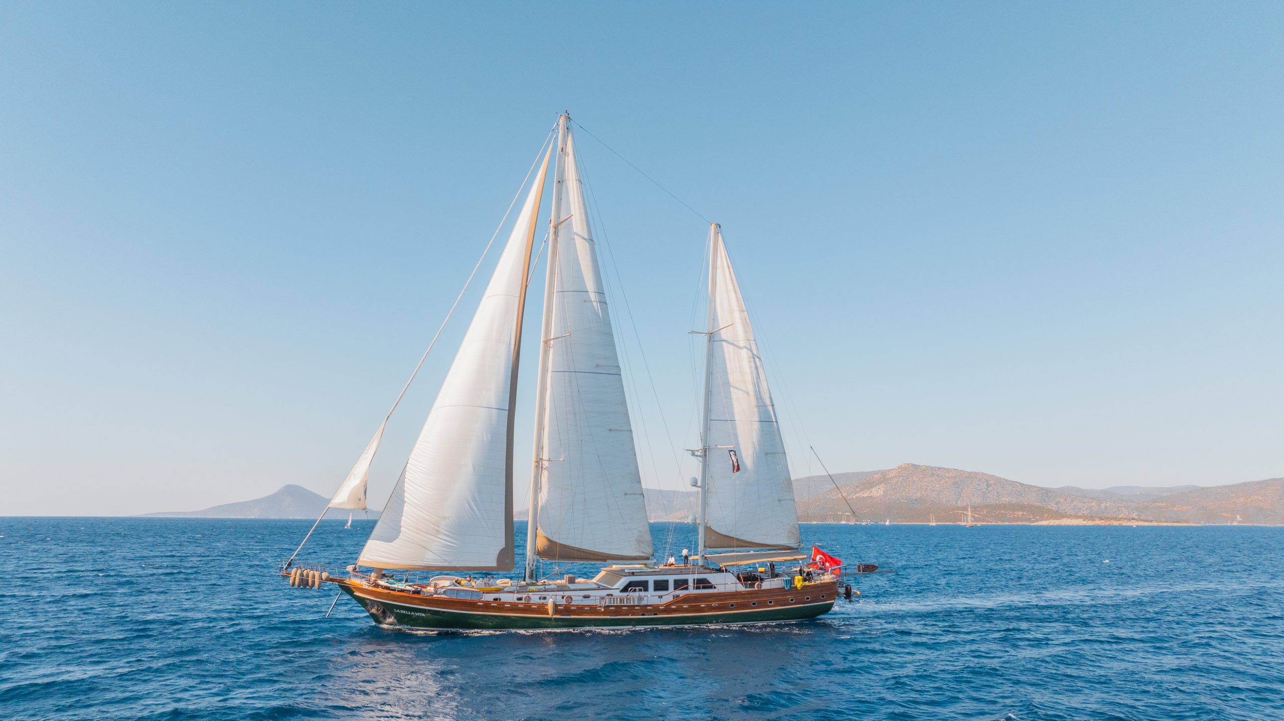 A large sailboat with white sails glides on calm blue water under a clear sky, with distant mountains visible in the background.