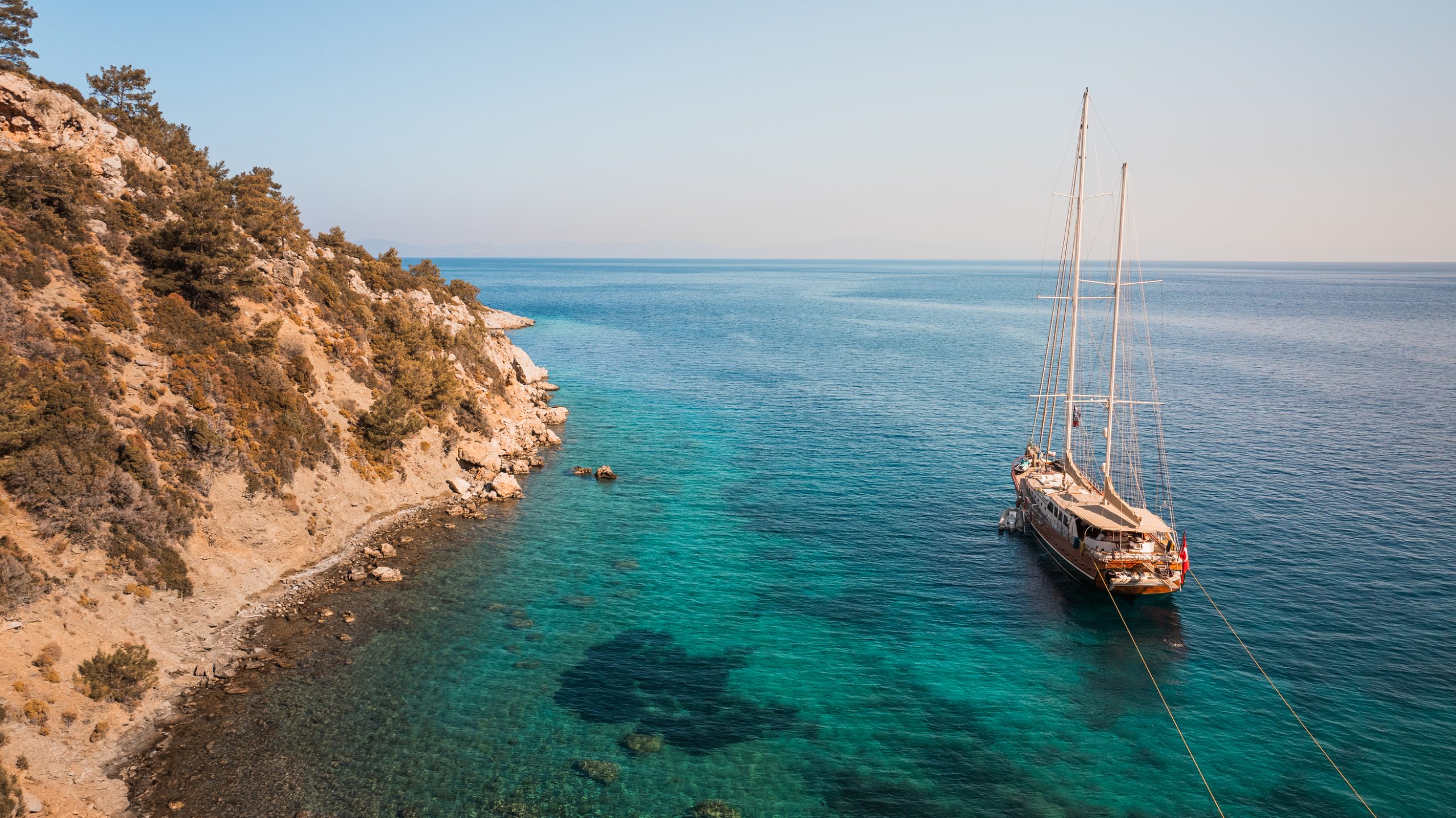 A sailboat is anchored near a rocky, tree-covered coastline with clear turquoise water, under a clear blue sky. The sea appears calm, and the shoreline curves gently to the left.