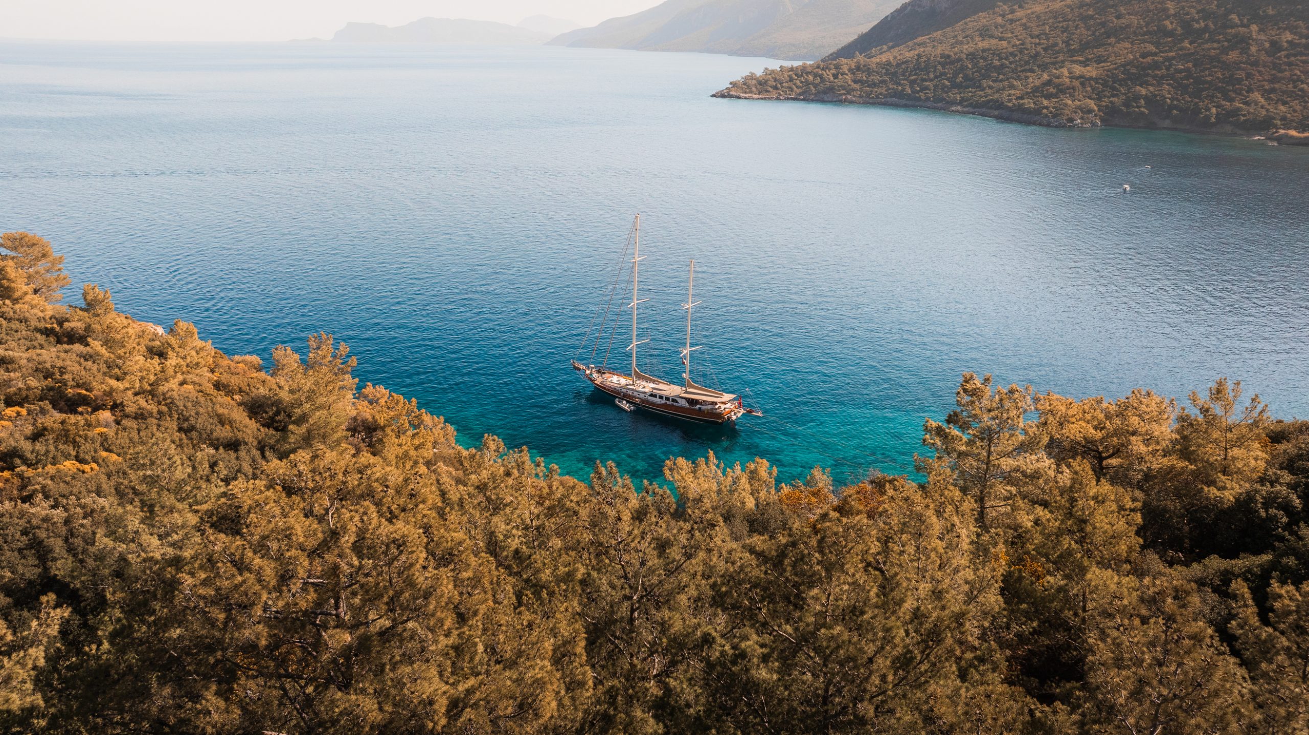 A sailboat with tall masts floats on calm blue water near the shore, surrounded by lush green trees and hills under a clear sky.