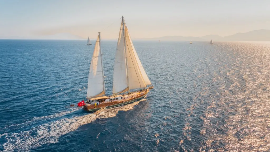 A sailboat with Turkish flag sails on a calm blue sea under a clear sky, with mountains and other sailboats visible in the distant background.
