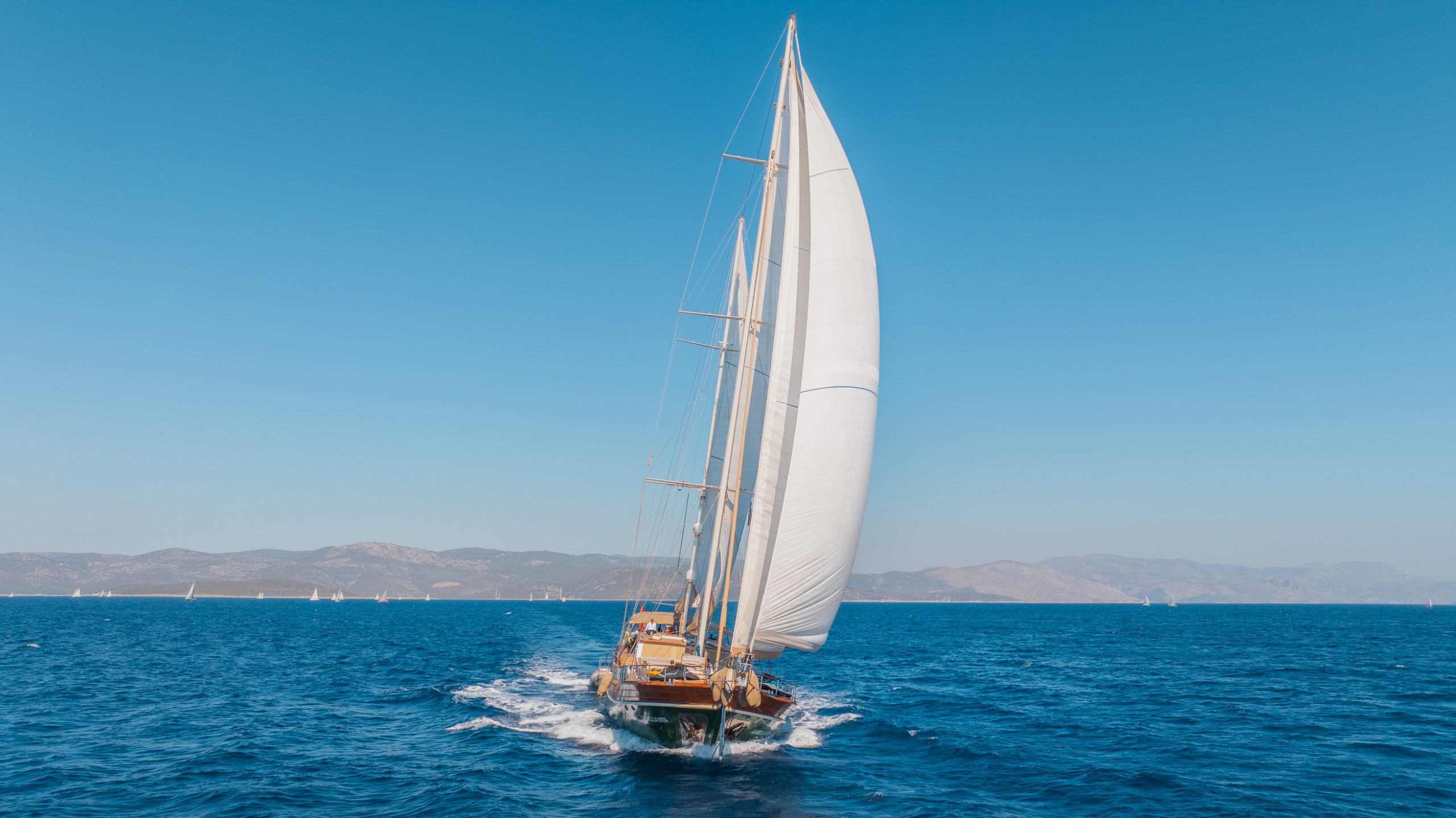 A large sailboat with white sails glides across deep blue water under a clear sky, with distant mountains and other sailboats visible on the horizon.