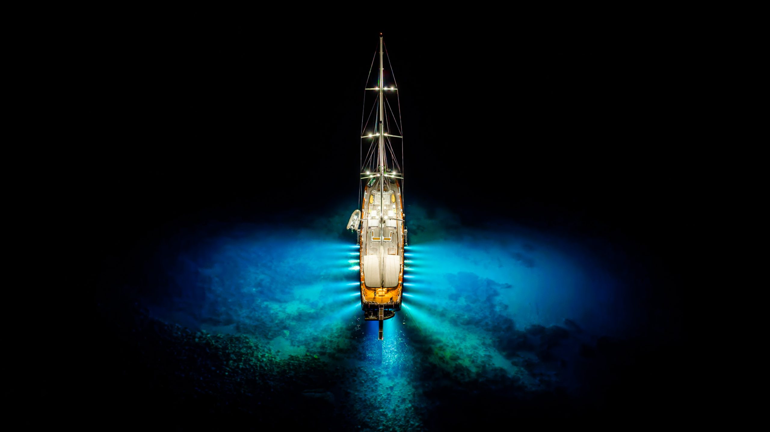 An illuminated sailboat is viewed from above at night, surrounded by glowing blue water, creating a striking contrast against the dark sea. The boats lights cast a circular glow on the water below.