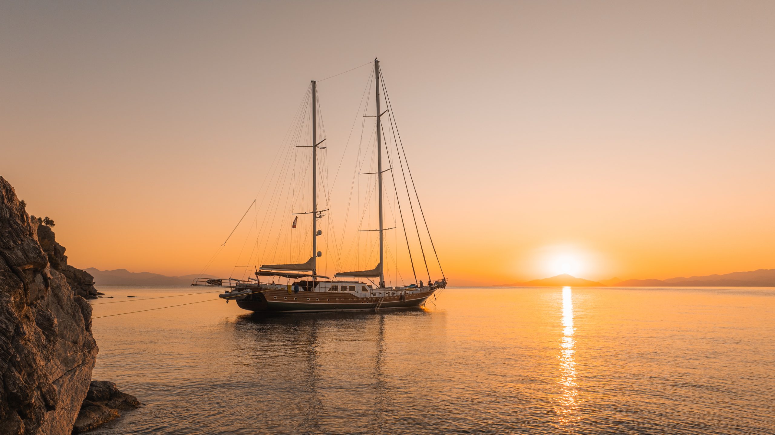 A sailboat anchored near a rocky shore at sunset, with calm water reflecting the orange glow of the sky and the sun setting behind distant mountains.
