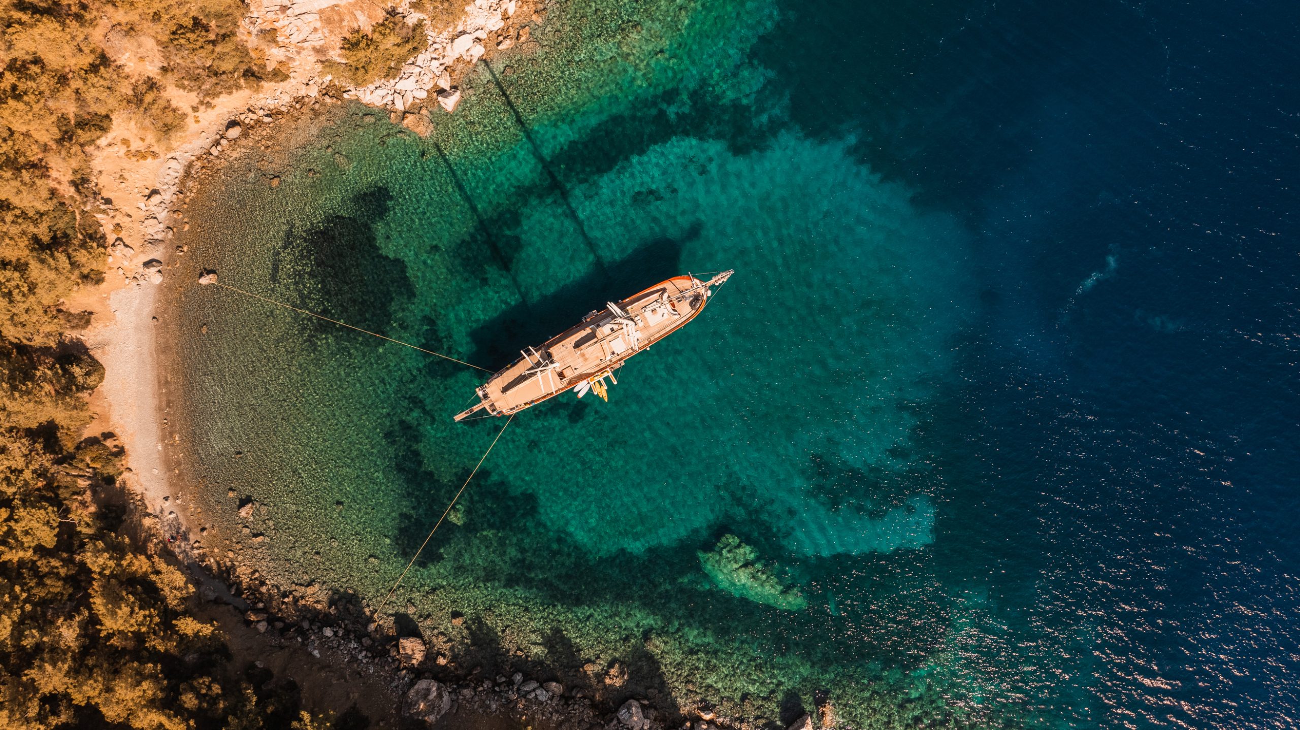 Aerial view of a boat anchored in clear turquoise water near a rocky shoreline, surrounded by trees and shallow reefs, with deep blue sea further out.
