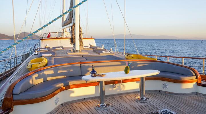 A yacht deck with curved cushioned seating, a white table set with drinks and snacks, and sun loungers. The boat is on calm blue water at sunset, with distant mountains visible on the horizon.