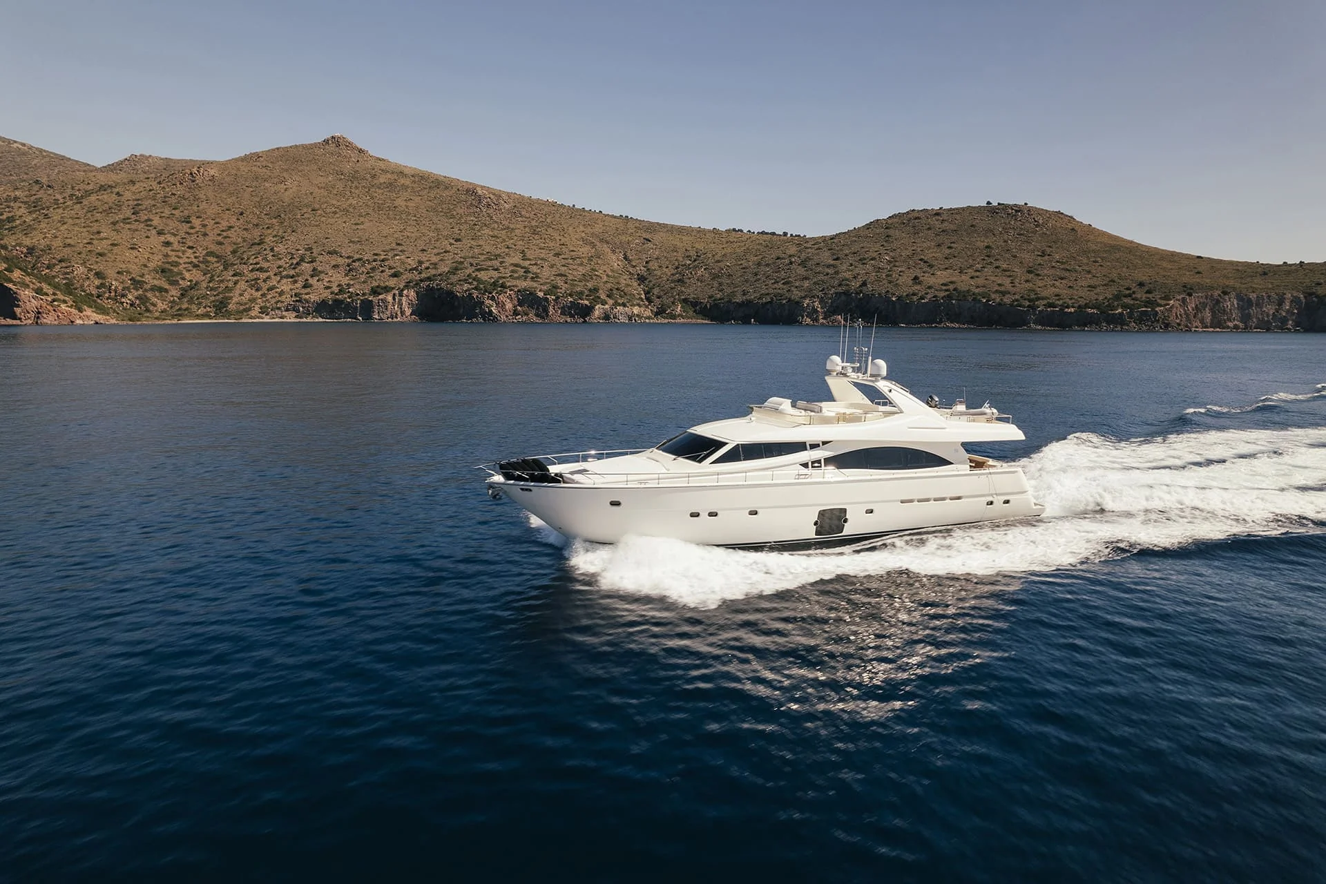 A white yacht cruises on calm blue water near a hilly coastline under a clear sky, creating a wake as it moves forward.