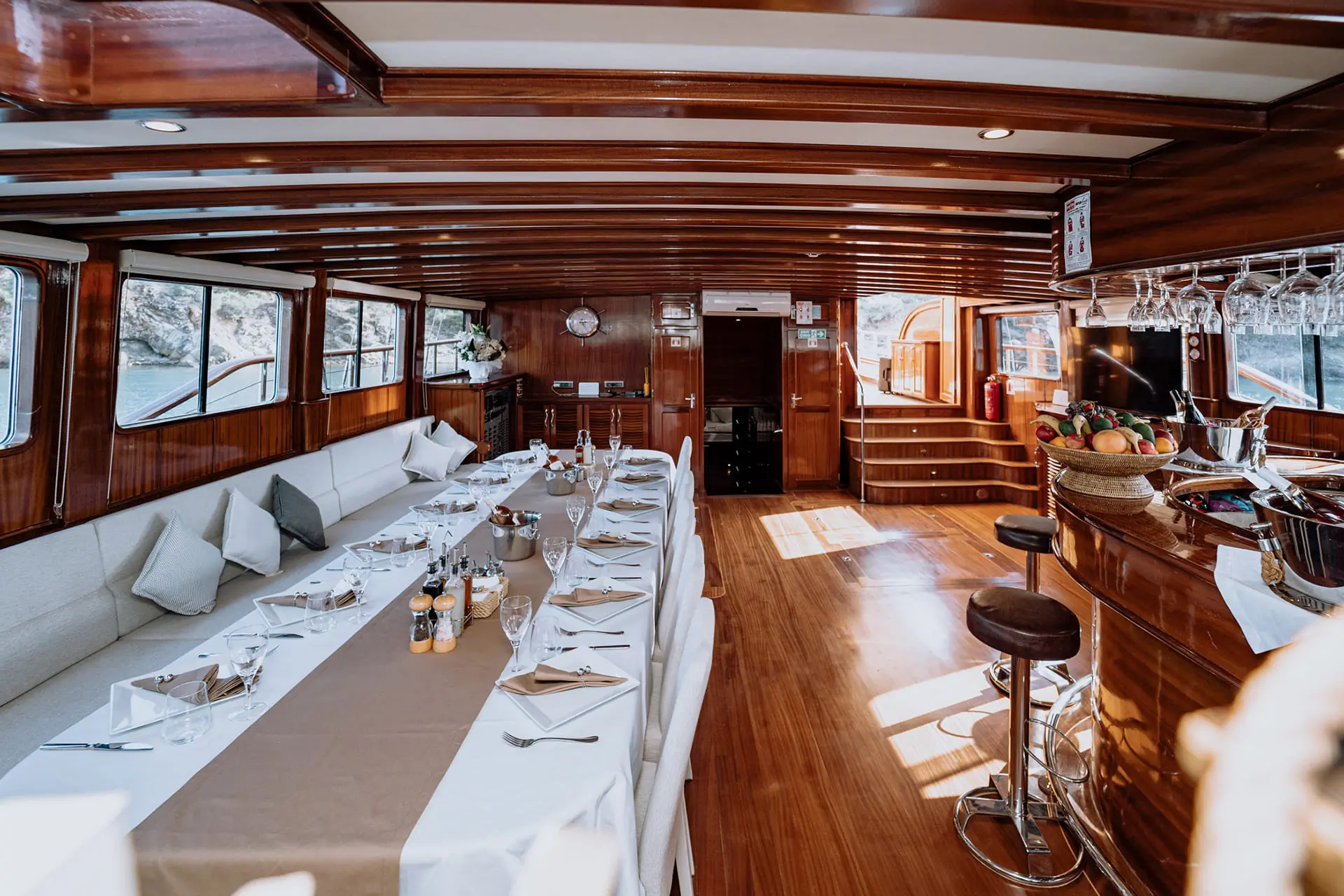 Elegant yacht interior with wooden walls and floors, a long dining table set for a meal, cushioned seating along the windows, bar area with stools, and stairs leading to another level. Natural light fills the space.