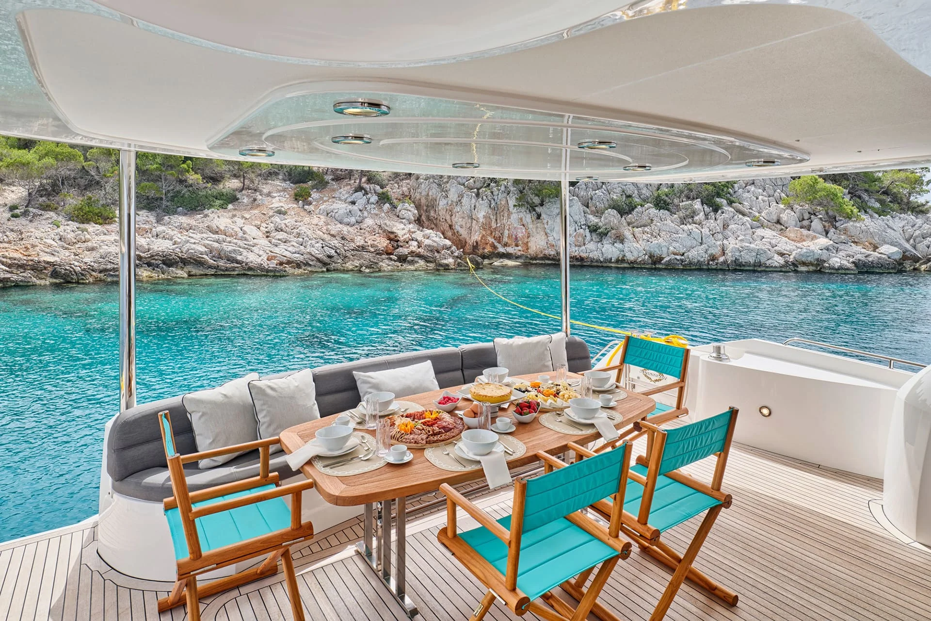 A dining table set for breakfast on a yacht deck overlooks clear turquoise water and rocky, tree-lined shore. Four blue chairs and a gray cushioned bench surround the table filled with food and drinks.