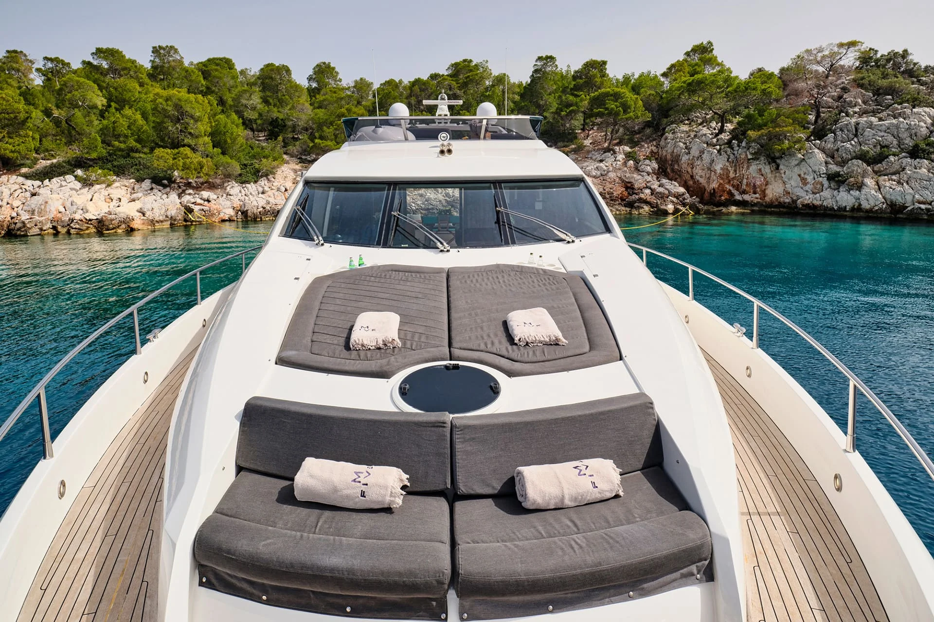 A luxury yacht deck with cushioned sunbeds and towels, anchored near rocky shoreline and green trees, overlooking clear blue water under a clear sky.
