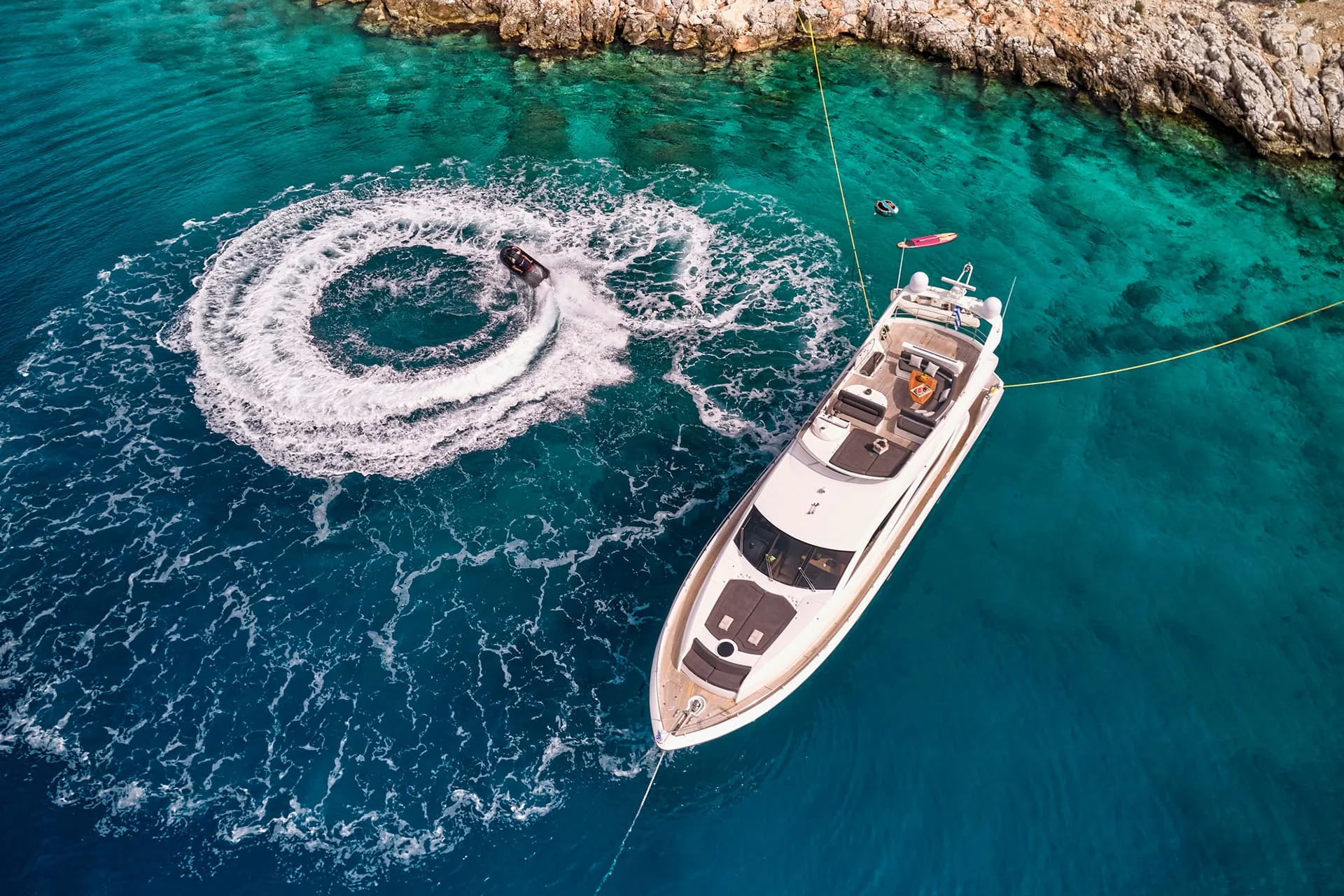 Aerial view of a yacht anchored in clear blue water near rocky shore, with a person riding a jet ski making circular waves nearby.