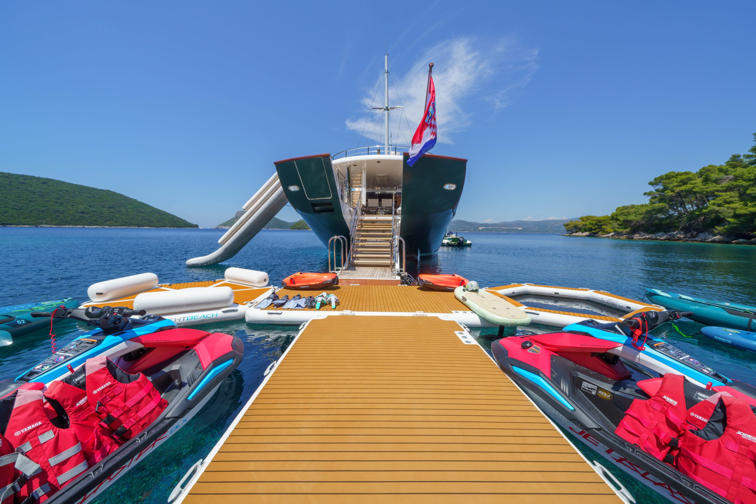 MAIA A luxury yacht with a slide is docked near a floating pier surrounded by jet skis, inflatable boats, and clear blue water, with green hills and a bright blue sky in the background.