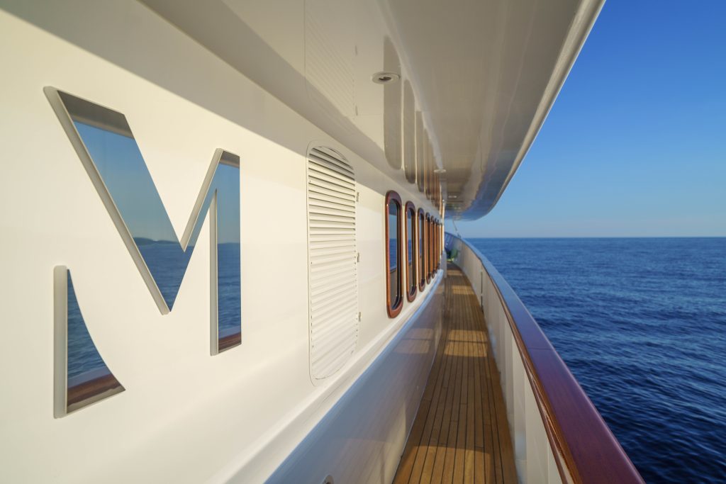 MAIA A close-up view of a yacht’s side deck with wooden flooring, circular windows, and a large “M” cutout in the white wall, overlooking calm blue ocean under a clear sky.