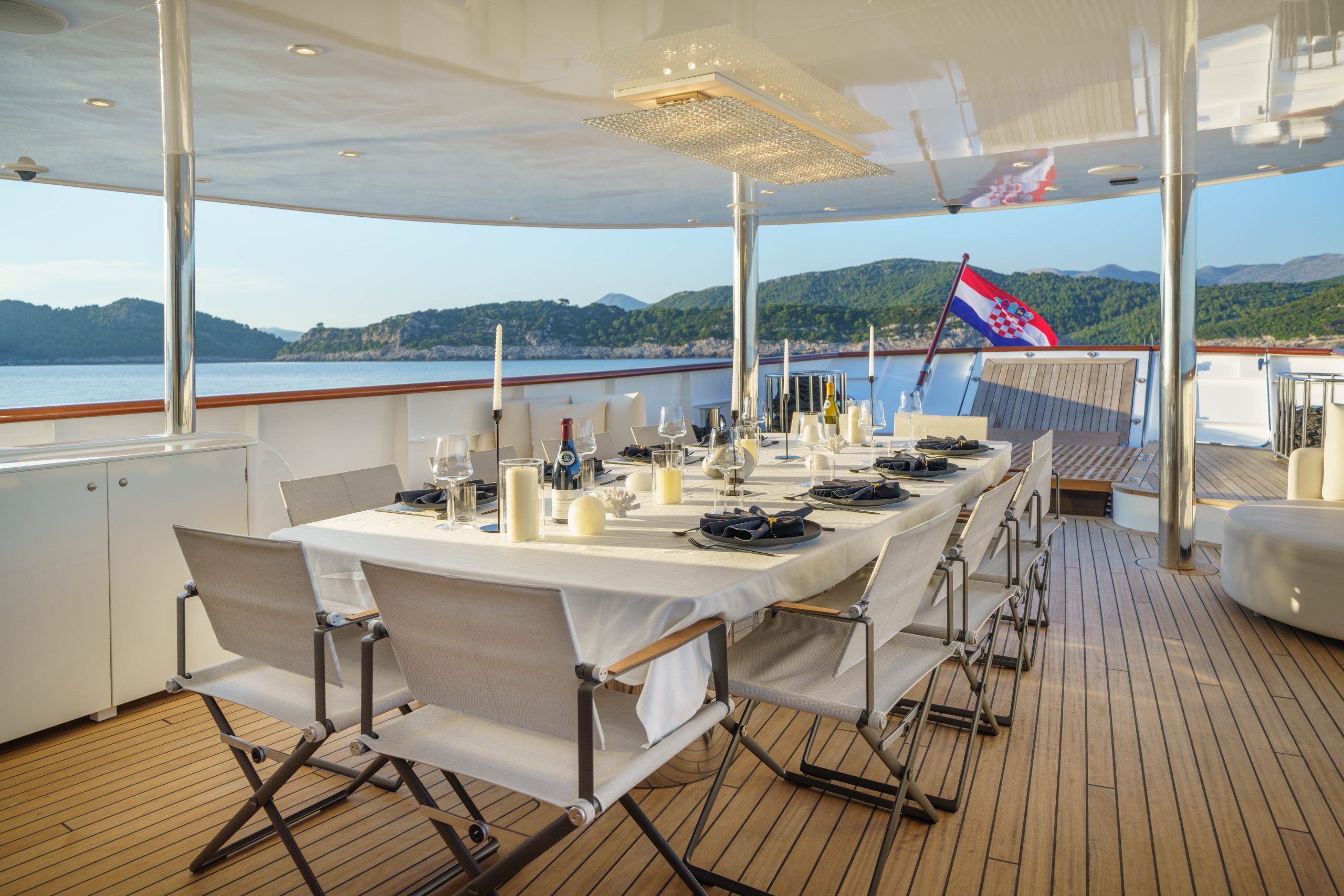 MAIA A dining table set for a meal is arranged on the deck of a yacht, with white chairs, candles, and tableware. The yacht overlooks calm blue water and a distant green shoreline, and a Croatian flag is visible at the back.