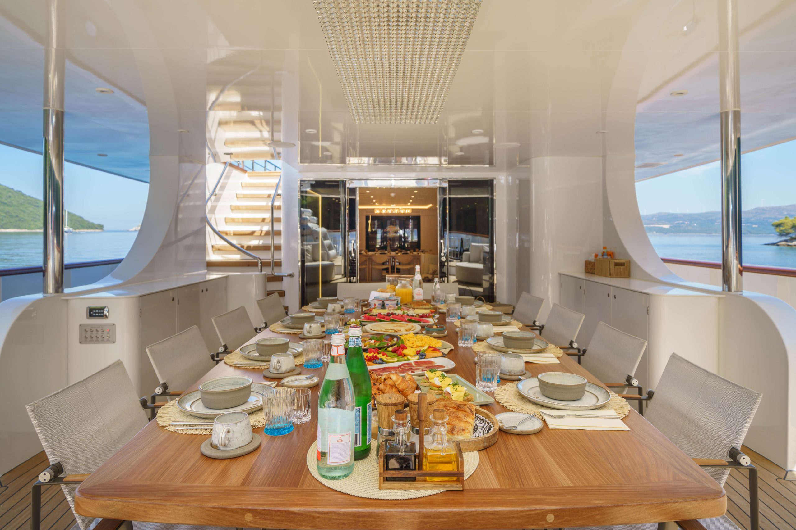 MAIA A luxurious yacht dining area features a large wooden table set for a meal with plates, glasses, bottled water, food platters, and bread. Sunlight streams in, and windows reveal views of the sea and distant green hills.