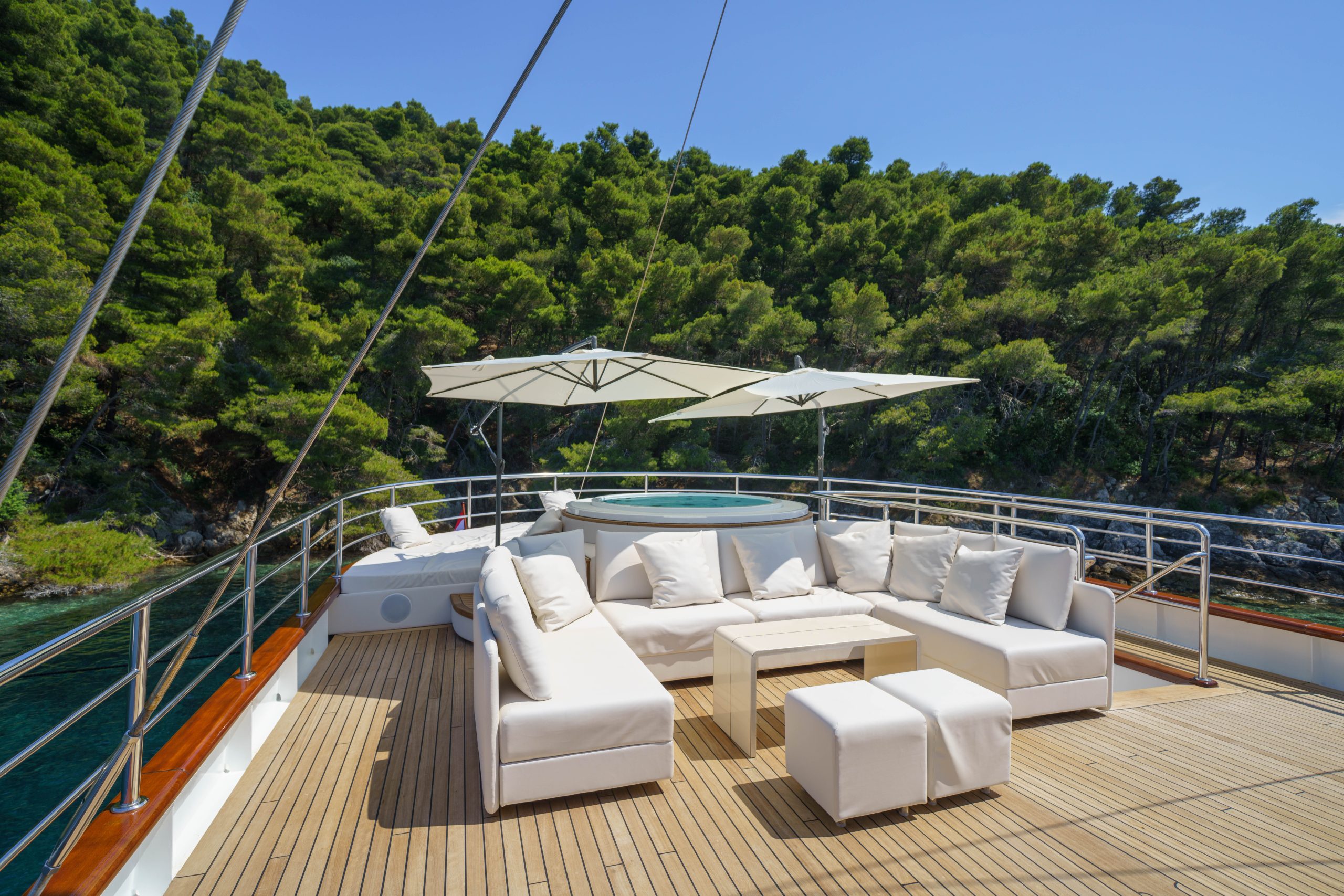 MAIA A luxurious yacht deck features white cushioned seating, a table, ottomans, umbrellas, and a hot tub, all set against a backdrop of green forest and clear blue sky.