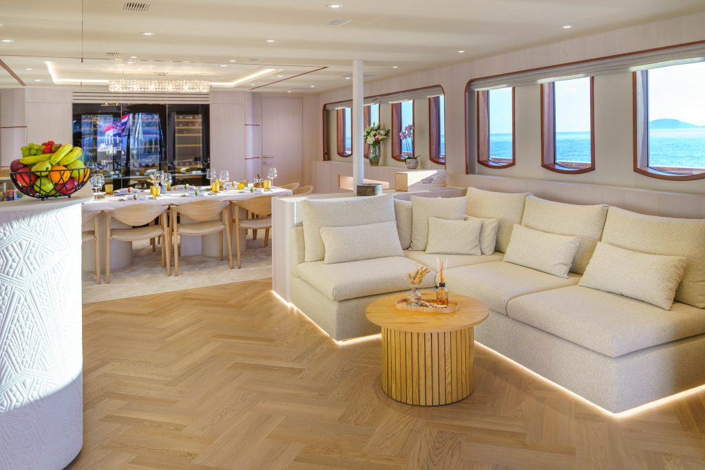 MAIA A luxurious yacht interior with a large cream-colored sectional sofa, a round wooden coffee table, a dining table set for a meal, fruit bowl, and large windows offering ocean views and natural light.