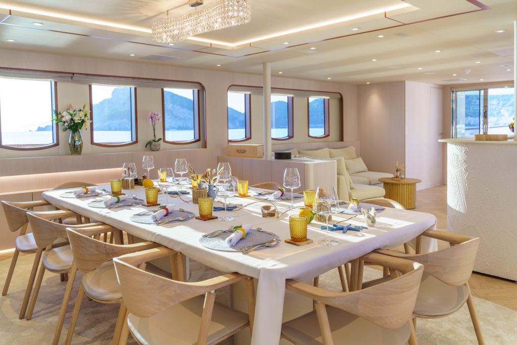 MAIA A luxurious yacht dining room with a set table, wooden chairs, elegant tableware, large windows with ocean and mountain views, soft lighting, and a lounge area in the background.