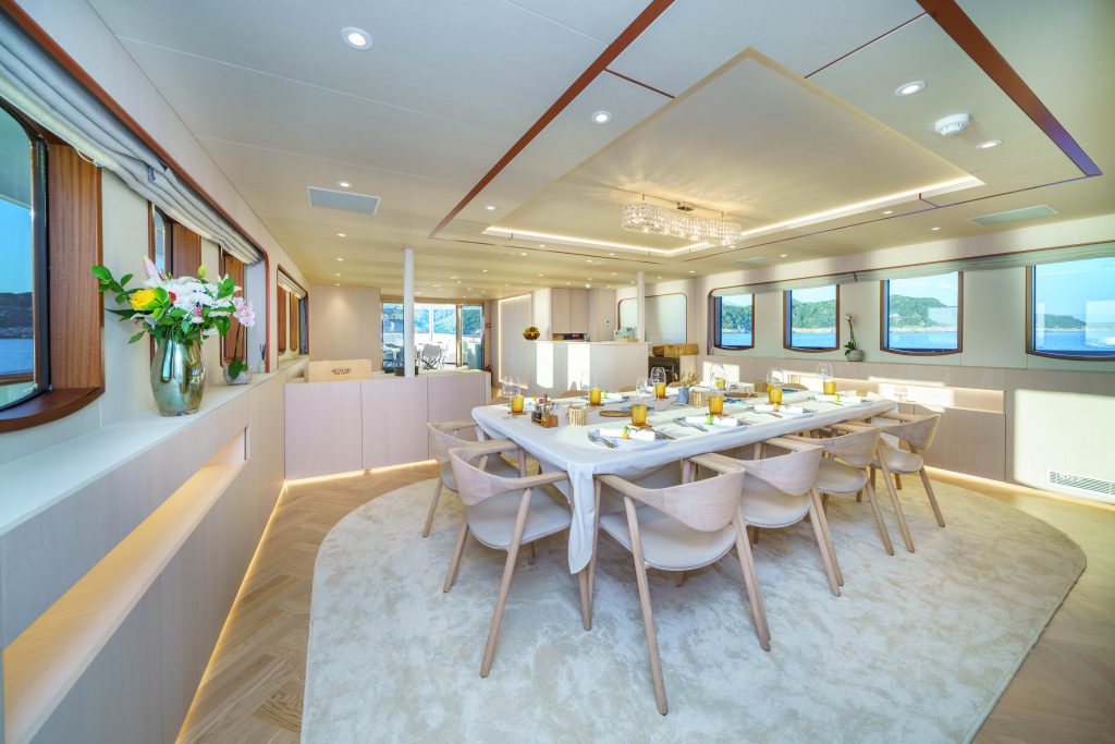 MAIA Elegant dining area on a yacht features a long table set for a meal, surrounded by light wooden chairs. Large windows offer ocean views, and fresh flowers decorate the bright, modern interior.