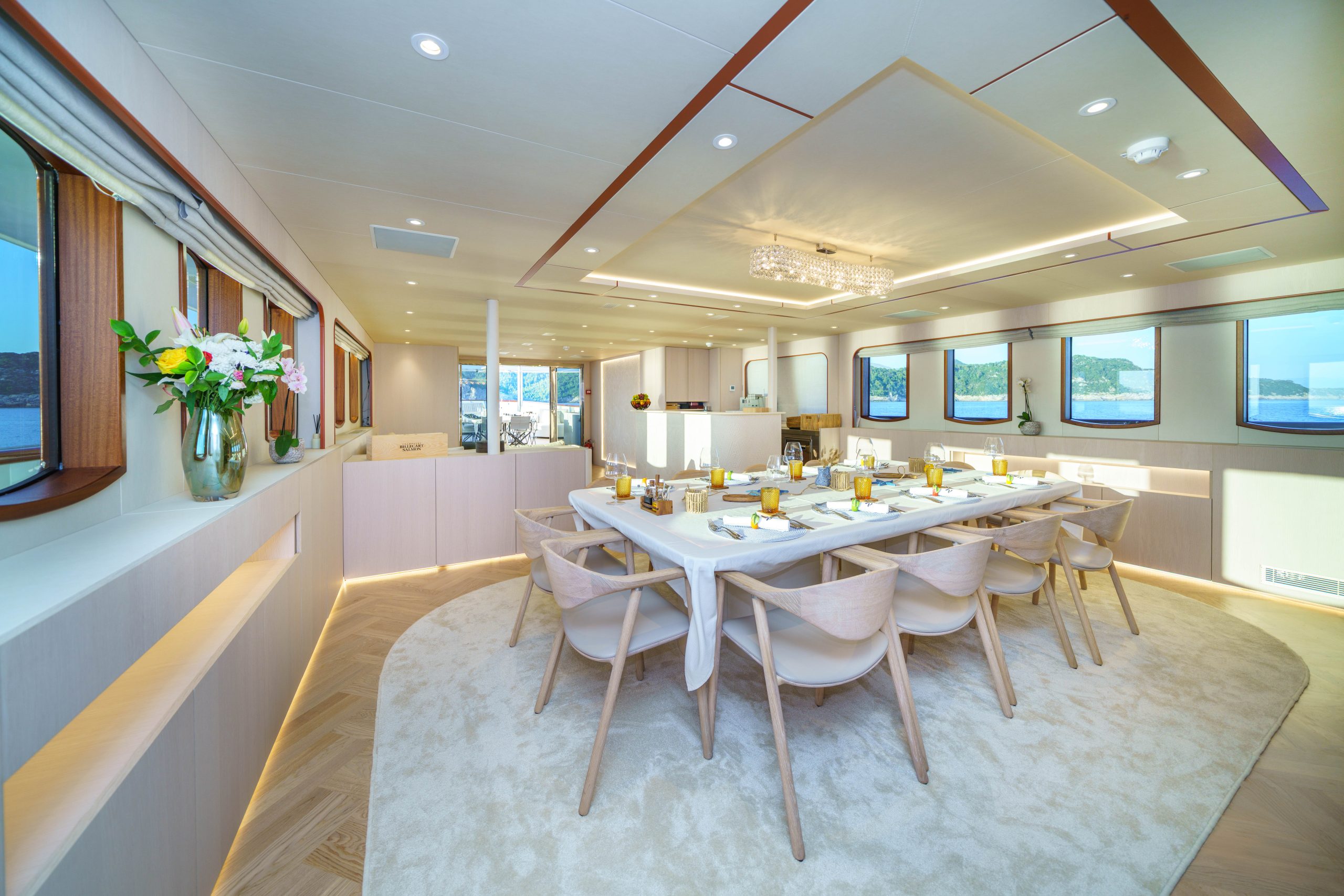 MAIA Elegant dining area on a yacht features a long table set for a meal, surrounded by light wooden chairs. Large windows offer ocean views, and fresh flowers decorate the bright, modern interior.