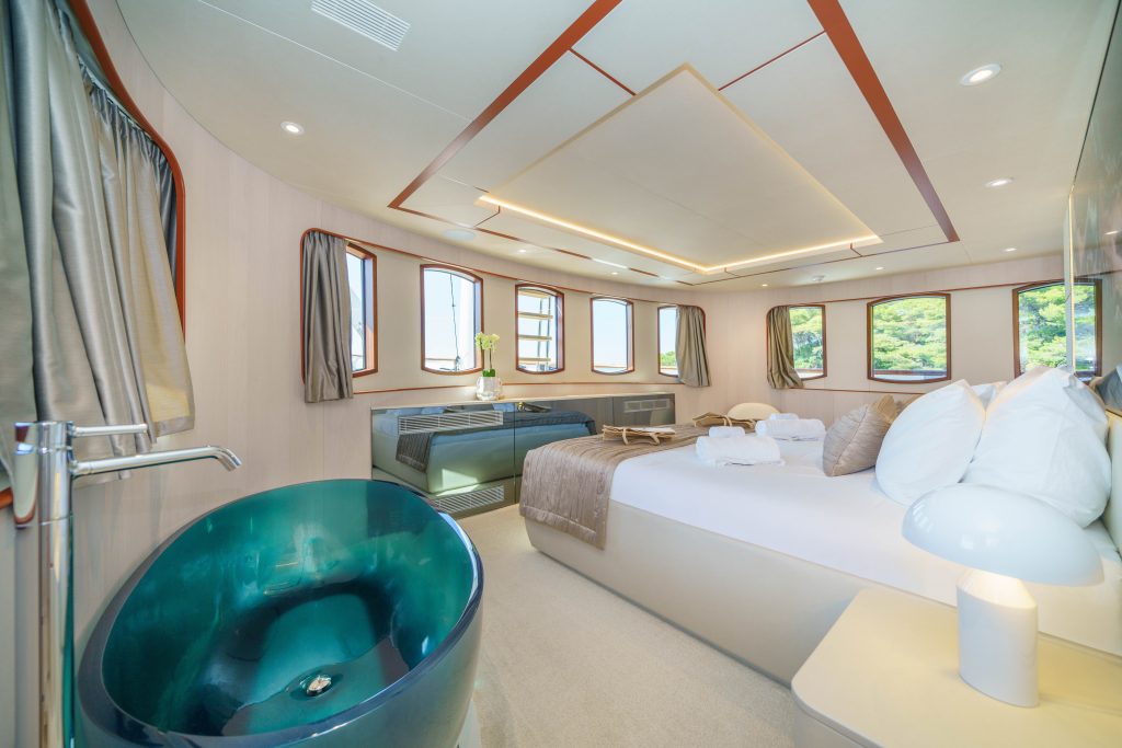 MAIA Spacious, modern yacht bedroom with large windows, a plush double bed, a sleek teal bathtub, a white lamp, a beige sofa, and light wood décor. Robes and towels are neatly placed on the bed.