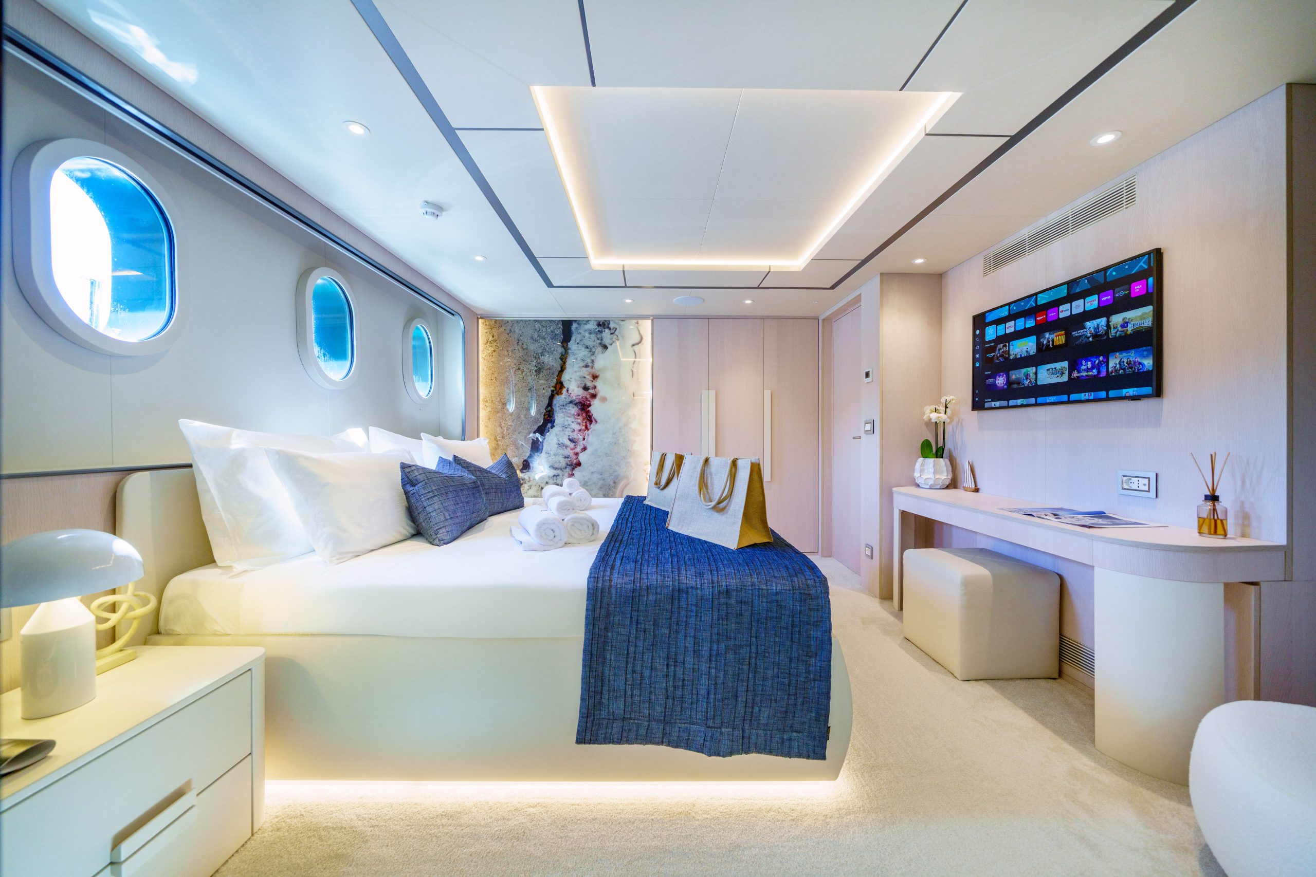 MAIA A luxurious yacht cabin with a large bed, blue blanket, round windows, modern decor, wall-mounted TV, and soft lighting, creating a spacious and elegant atmosphere.