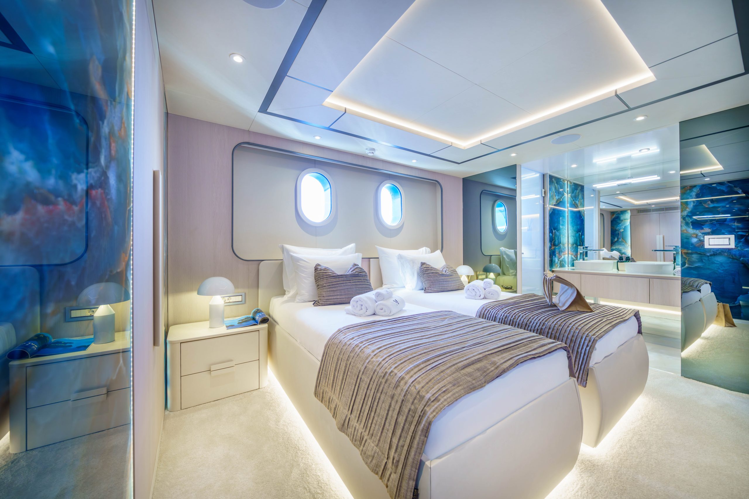 MAIA Modern yacht bedroom with two single beds, striped throws, and rolled towels. Two round windows bring in natural light. The room features sleek lighting, blue accent walls, and an open view of a connected bathroom.