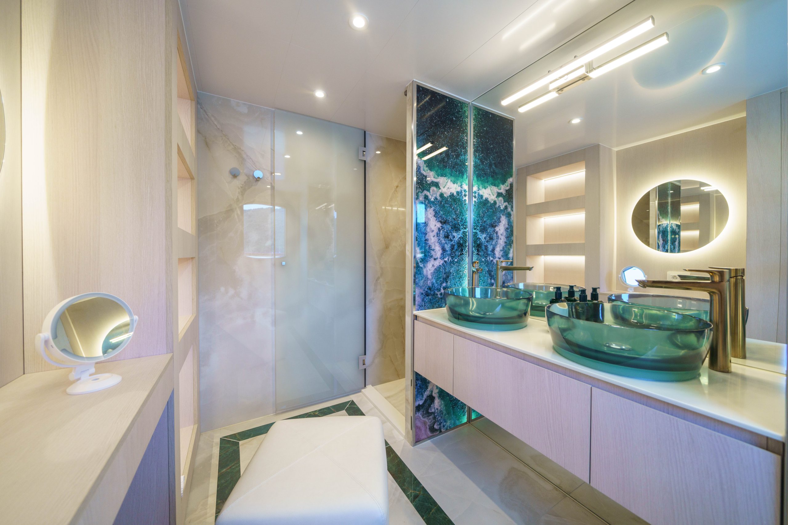 MAIA Modern bathroom with glass shower, double green glass sinks, large mirror, illuminated shelves, and a vanity stool. Marble accents and elegant lighting create a luxurious, contemporary atmosphere.