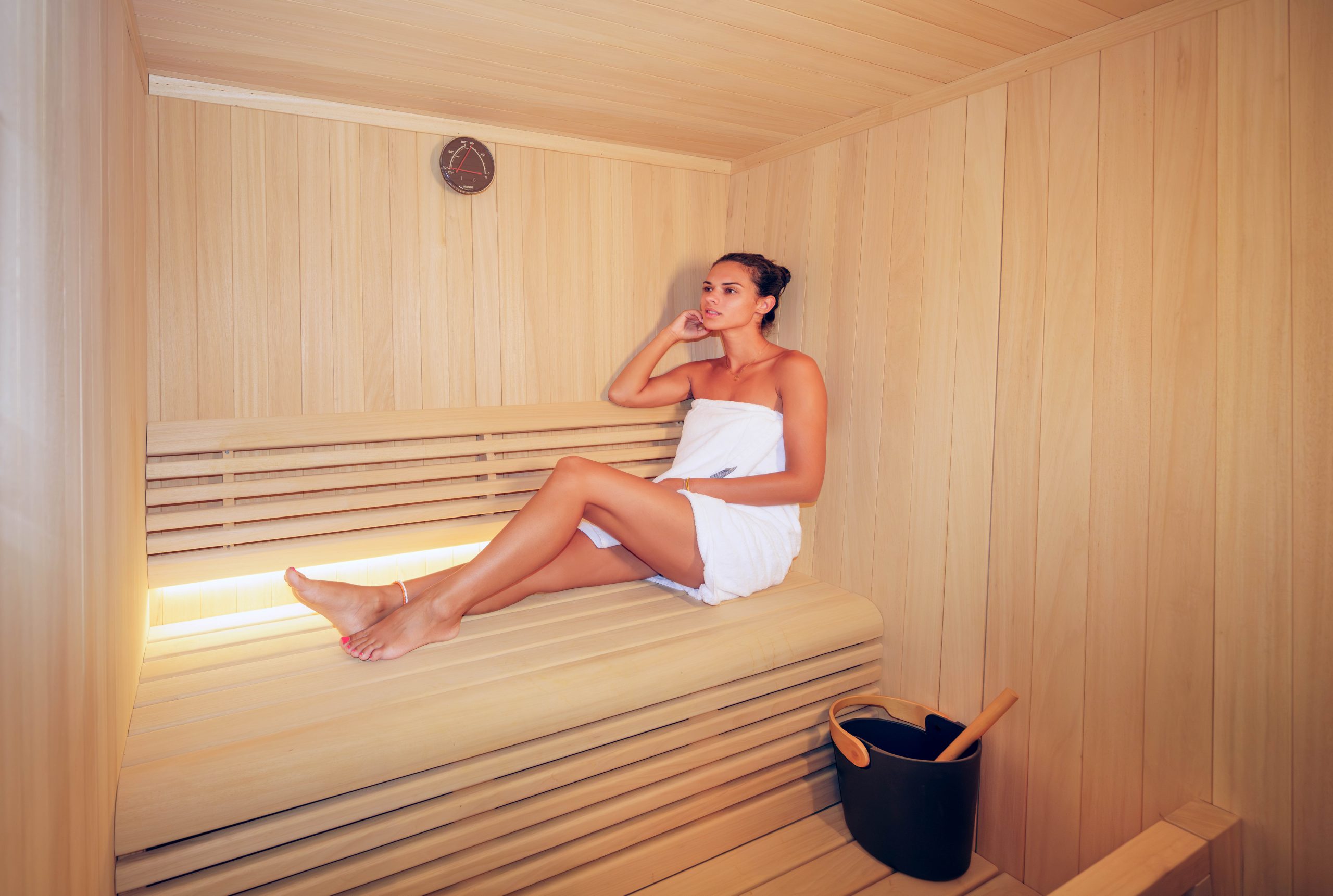 MAIA A woman wrapped in a white towel sits relaxing inside a wooden sauna. A small black bucket with a ladle is on the floor, and a clock is mounted on the wall above her. The light is warm and soft.