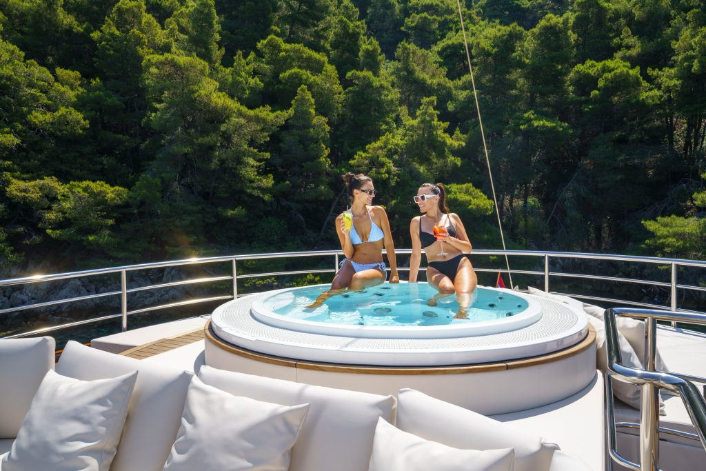 MAIA Two women in swimsuits sit in a hot tub on the deck of a yacht, enjoying drinks and talking, with lush green trees and hills visible in the background.