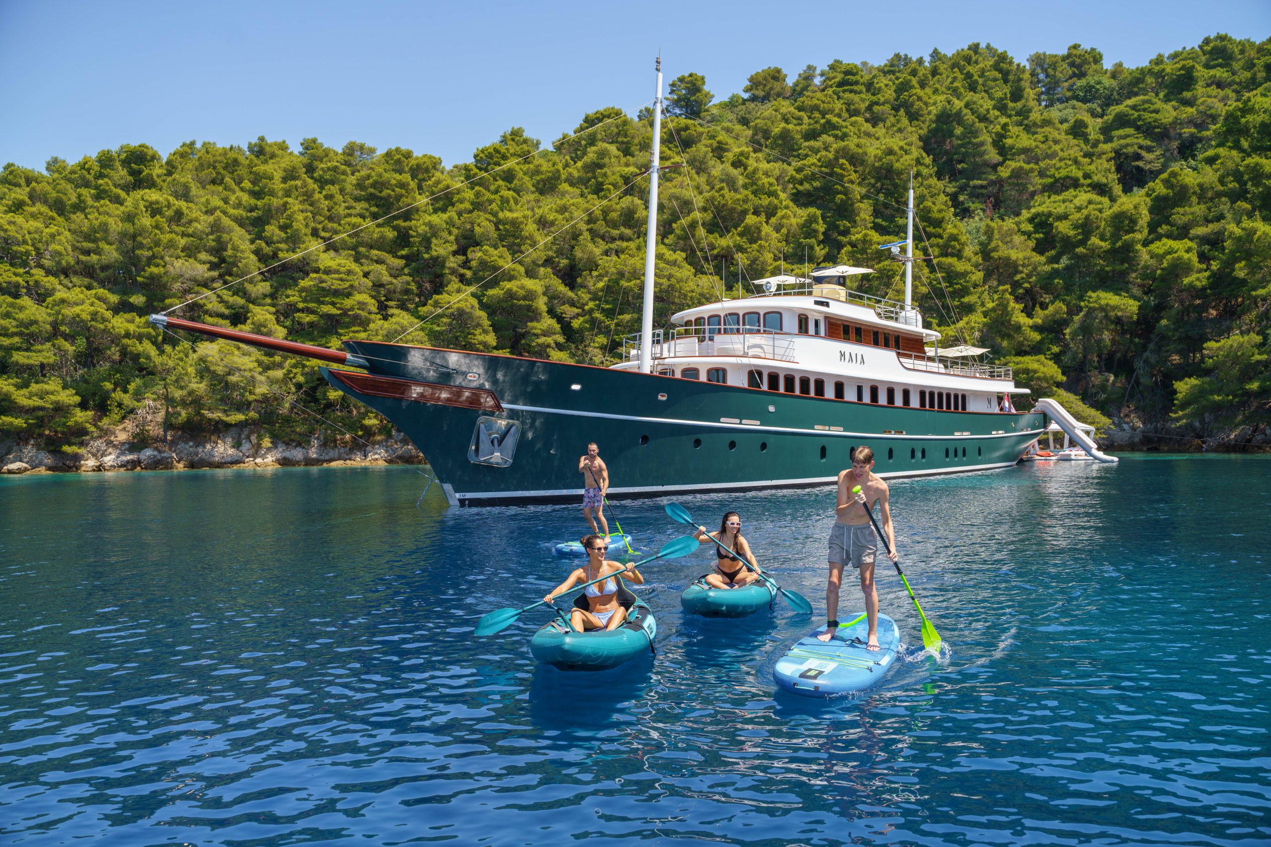 MAIA A luxury yacht is anchored near a forested shoreline. Three people paddle on blue boards in the water while another stands on the deck. The water is clear and calm, reflecting the greenery and yacht.