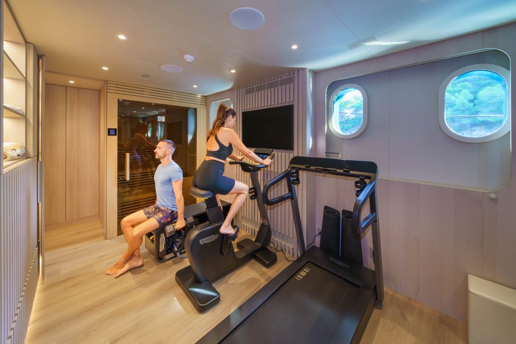MAIA A man and a woman exercise in a modern gym with wood floors and natural light. The man sits on a bench while the woman uses a stationary bike. Treadmill and porthole windows are visible. Sauna room can be seen in the background.