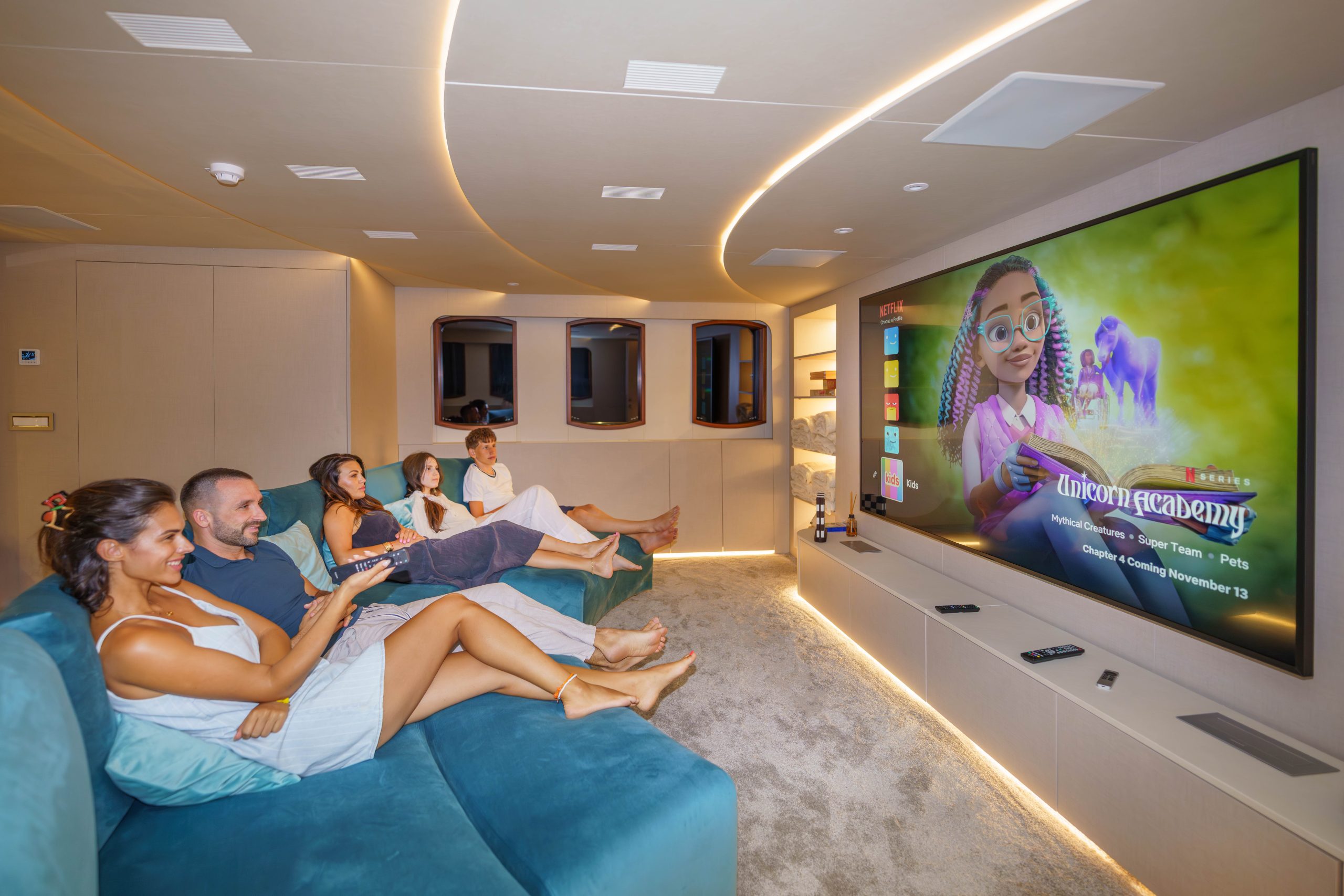 MAIA Four people relax on a blue sectional sofa in a modern home theater, watching a large screen displaying the Netflix show “Unicorn Academy.” The room is softly lit, cozy, and stylishly designed.