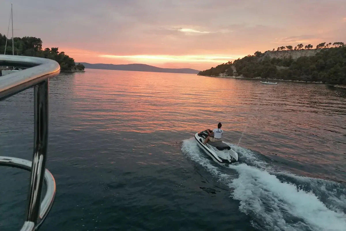 Person riding a jet ski on calm water during sunset, surrounded by trees and hills, with a partly cloudy sky and warm colors reflected on the water.