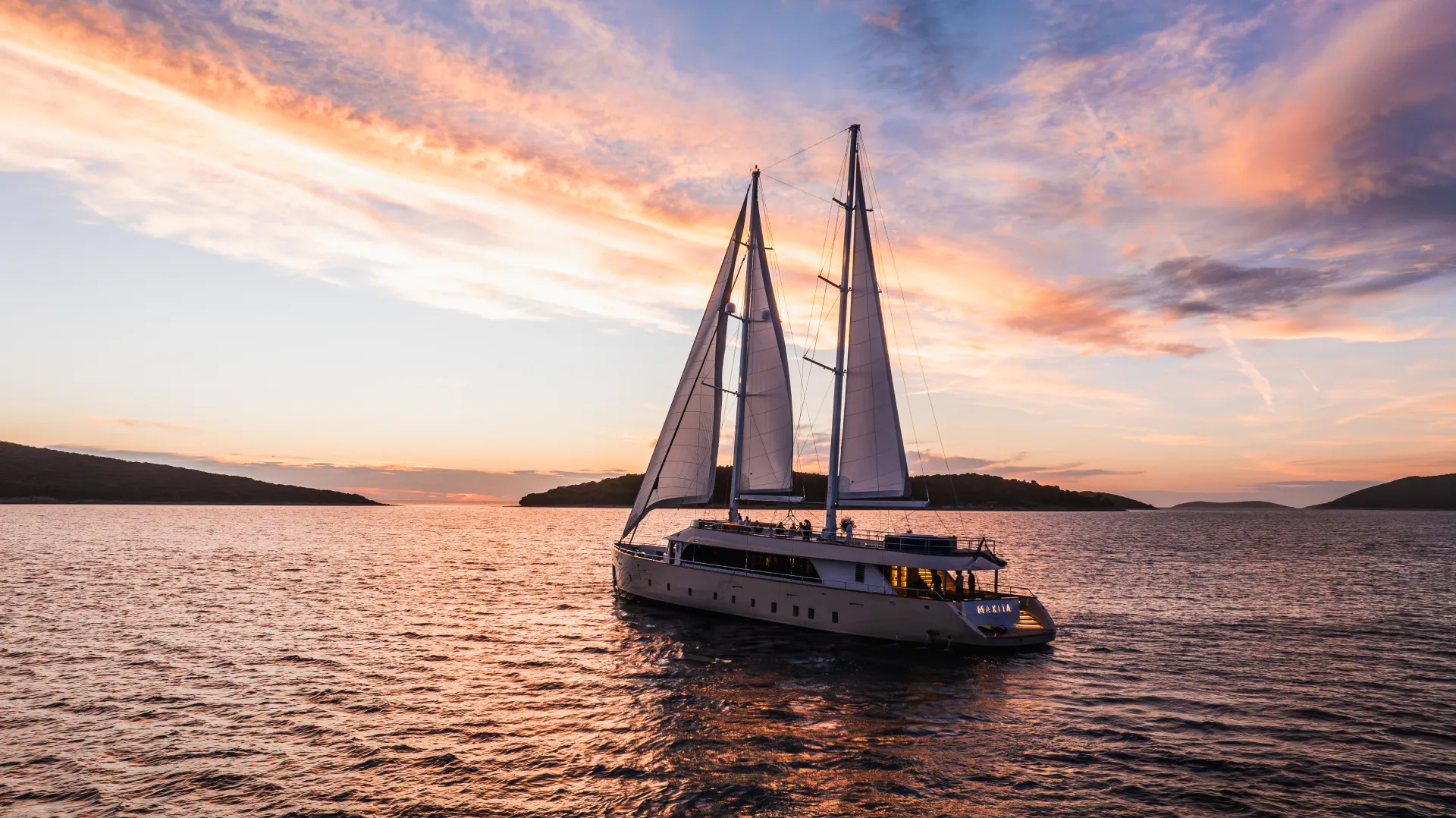 MAXITA A large sailboat with two tall masts sails on calm water at sunset, with vibrant orange, pink, and purple clouds filling the sky and distant hills on the horizon.