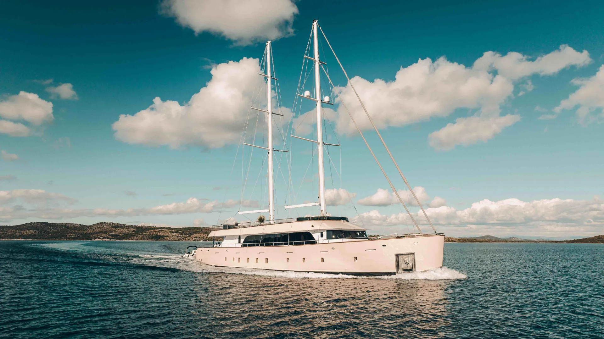 A large, modern yacht with two tall masts sails on calm blue water under a partly cloudy sky, with distant hills visible on the horizon.