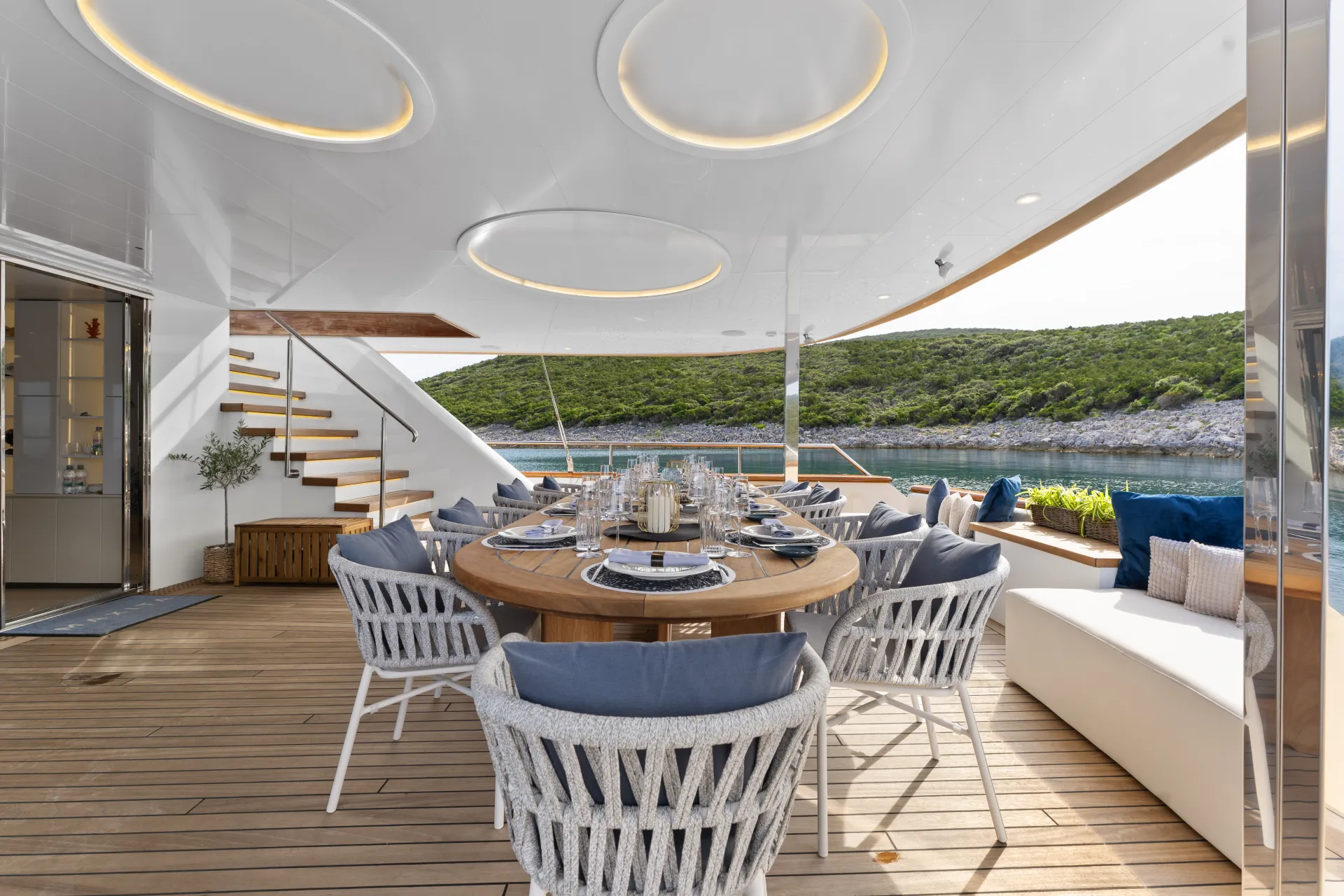 MAXITA Outdoor dining area on a yacht with a round table set for six, cushioned chairs, and modern decor. Overhead lights, nearby stairs, and water with green hills in the background create a luxurious seaside atmosphere.