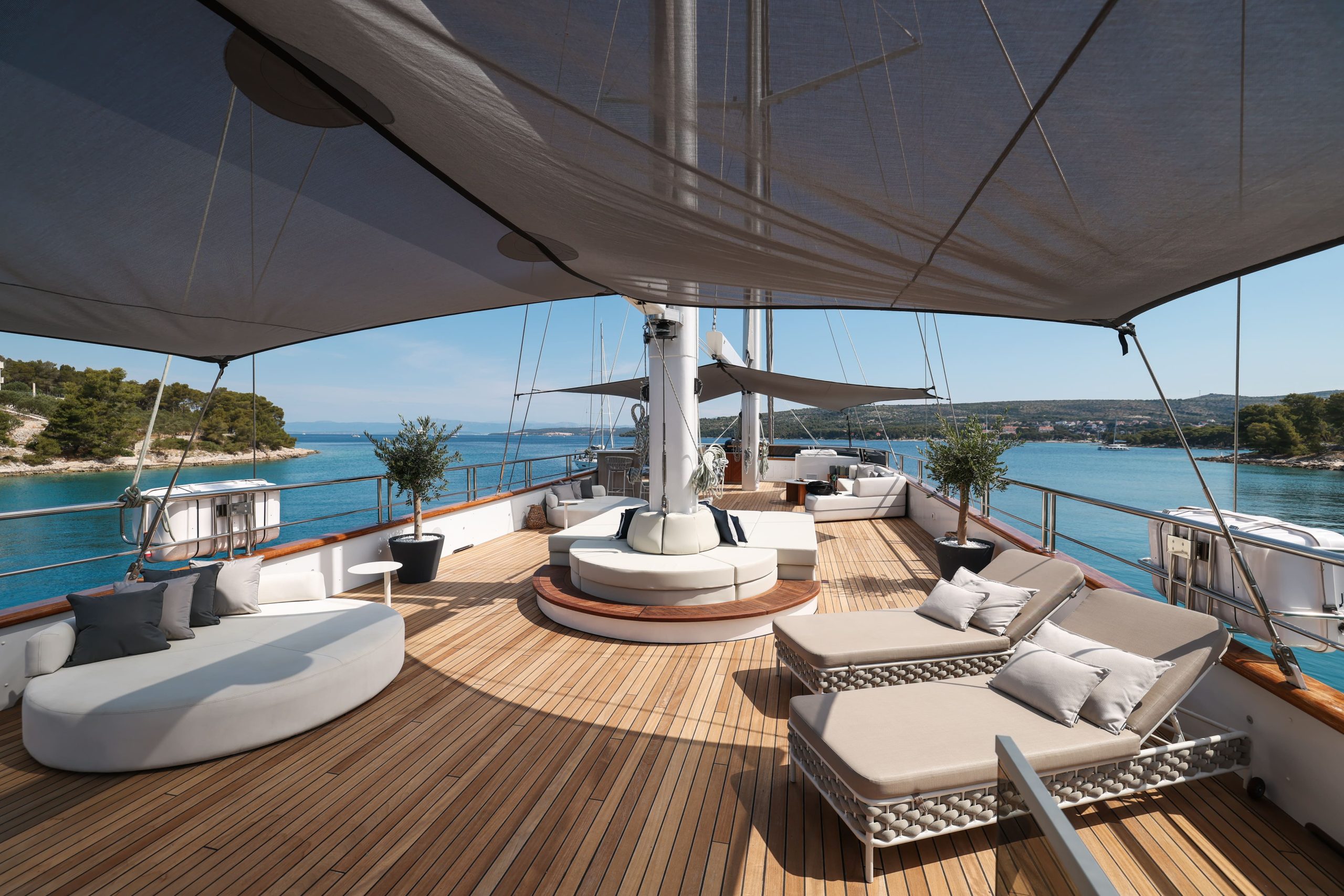 The deck of a yacht features multiple seating areas, including circular lounges and sun loungers, shaded by an overhead canopy, with a view of a blue sea and coastline in the distance.