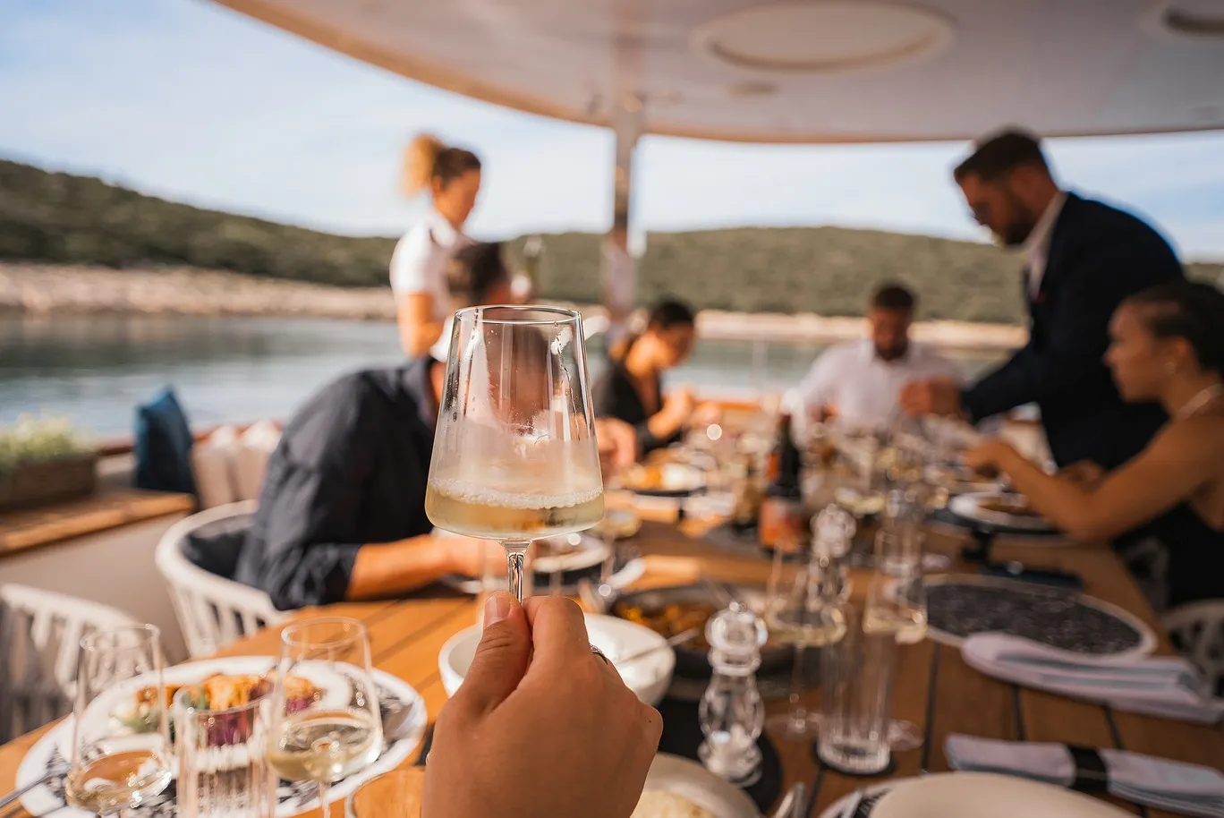 A hand holds up a glass of white wine toward a table set for a meal on a boat, with several people dining and scenic water and hills in the background.