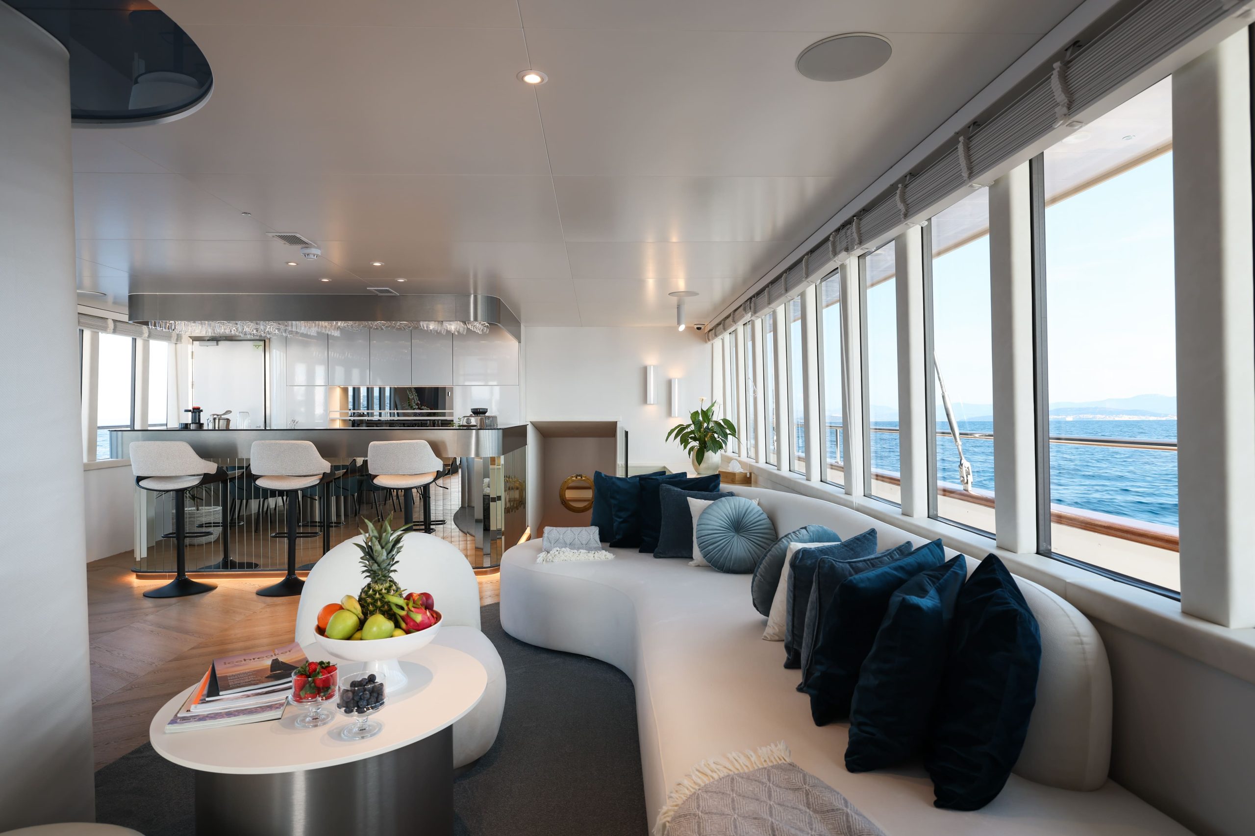 Interior of a yacht lounge featuring a curved white sofa with blue pillows, a coffee table with a bowl of fruit, and a bar area in the background overlooking the ocean.