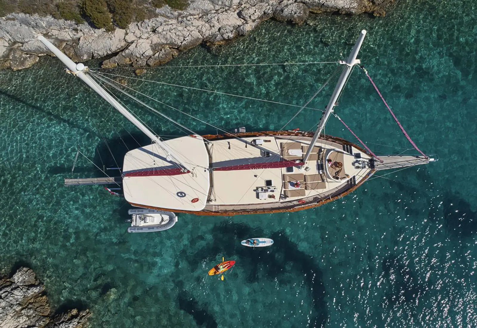 Aerial view of a sailboat anchored near a rocky shore in clear turquoise water, with a small inflatable boat, a paddleboard, and a kayak nearby.