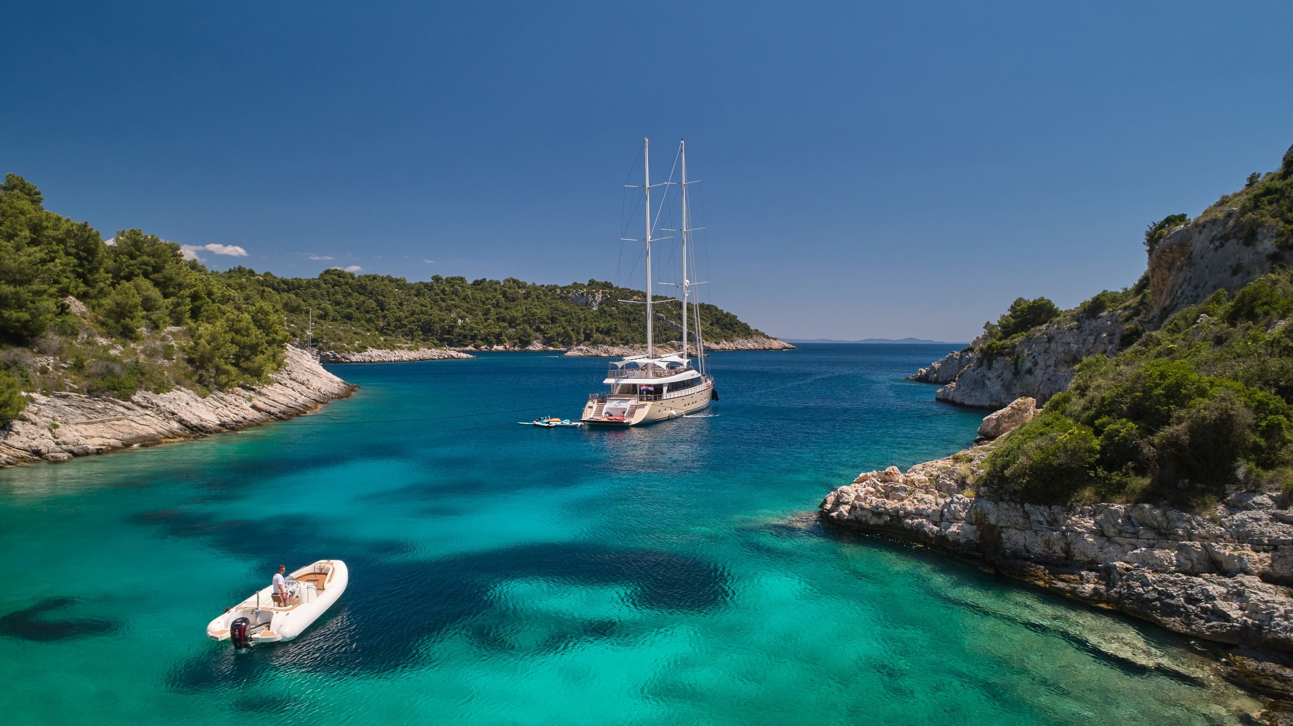 MARALLURE A large sailboat and a small motorboat float in a clear, turquoise bay surrounded by rocky, tree-covered hills under a bright blue sky. Two people relax in the motorboat.