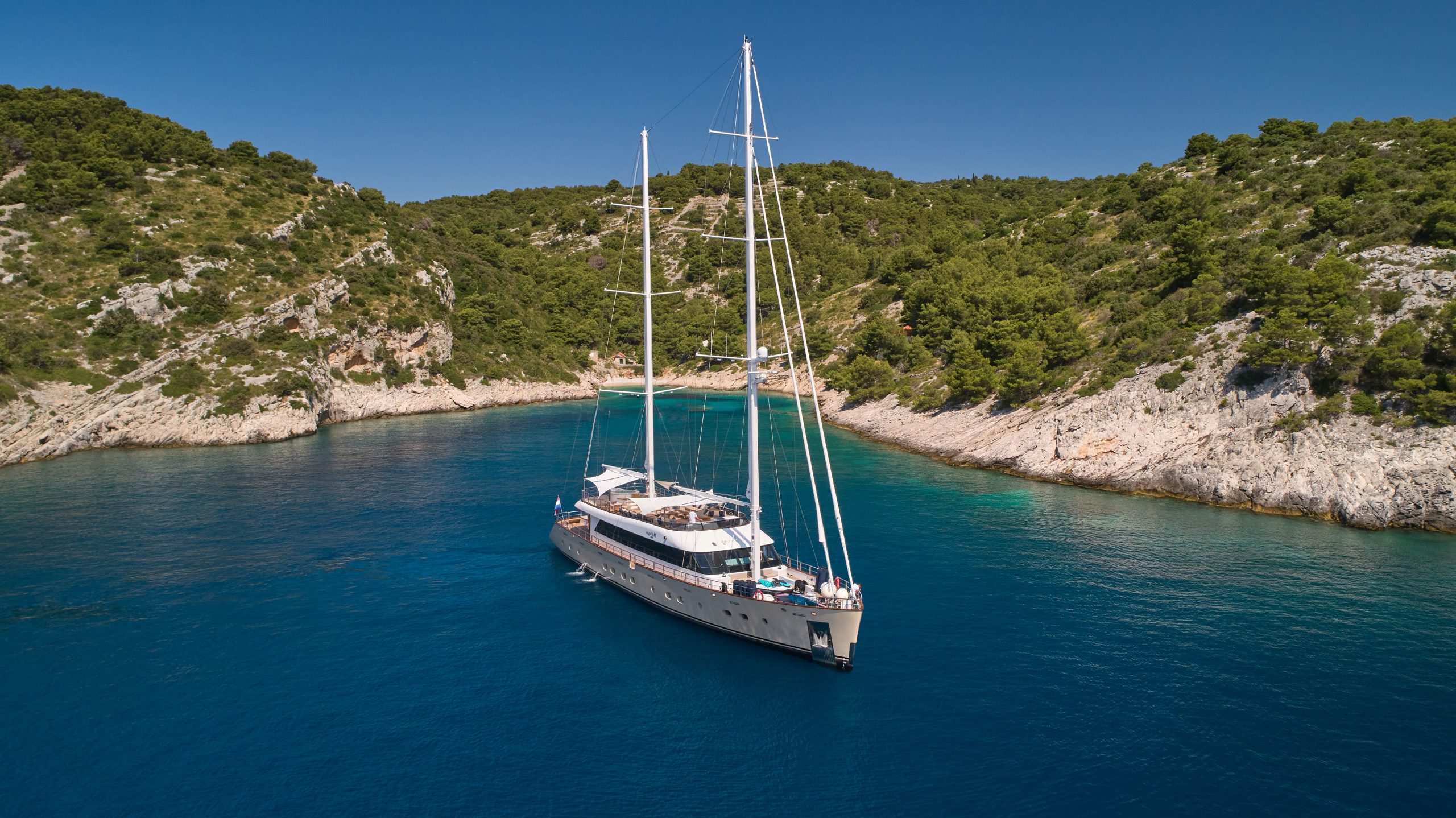 MARALLURE A large white yacht is anchored in a secluded blue bay surrounded by rocky, forested hills under a clear sky.