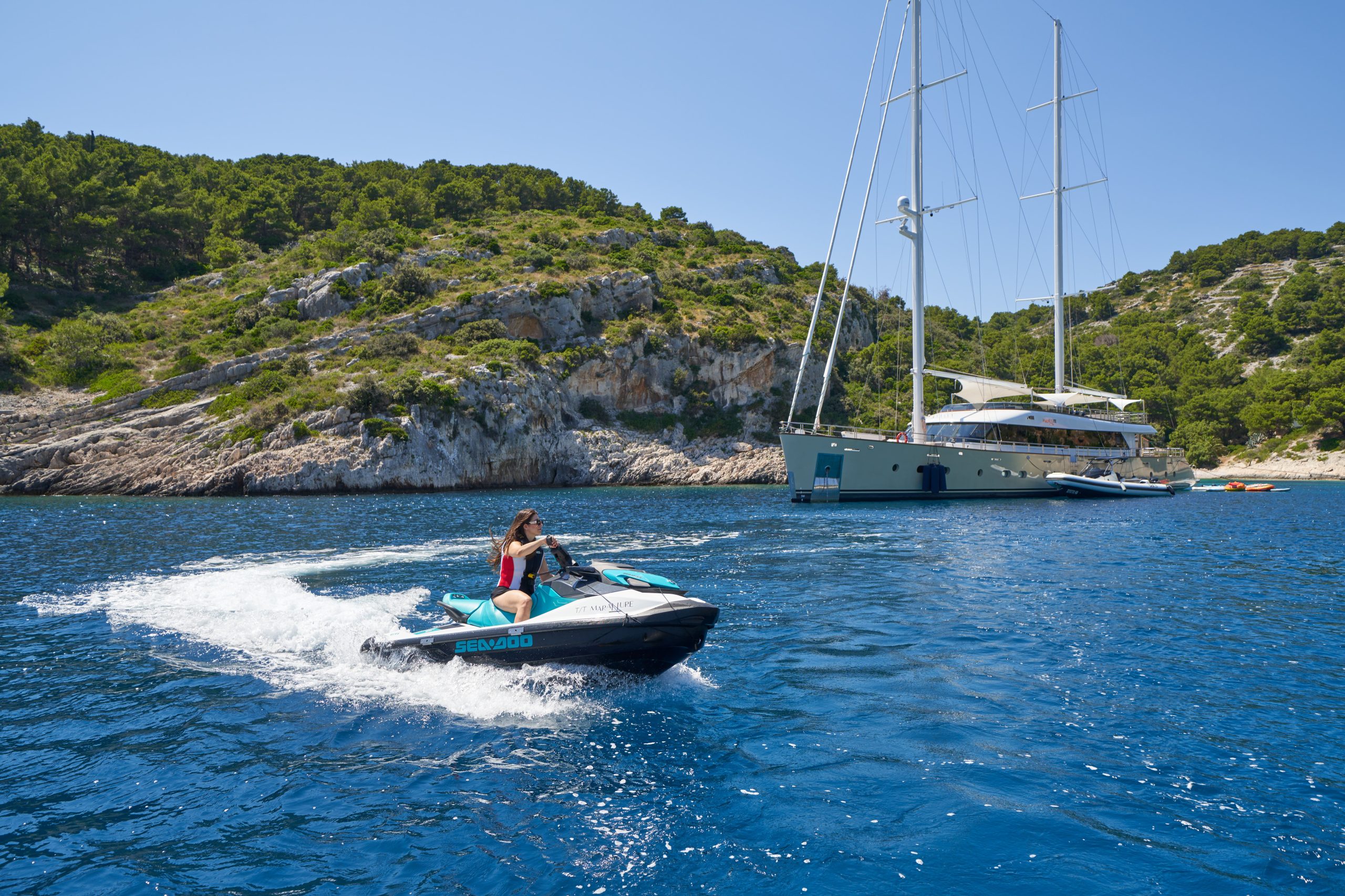MARALLURE A person rides a jet ski across blue water near a large sailboat anchored by a rocky, green-covered shoreline under a clear, sunny sky.