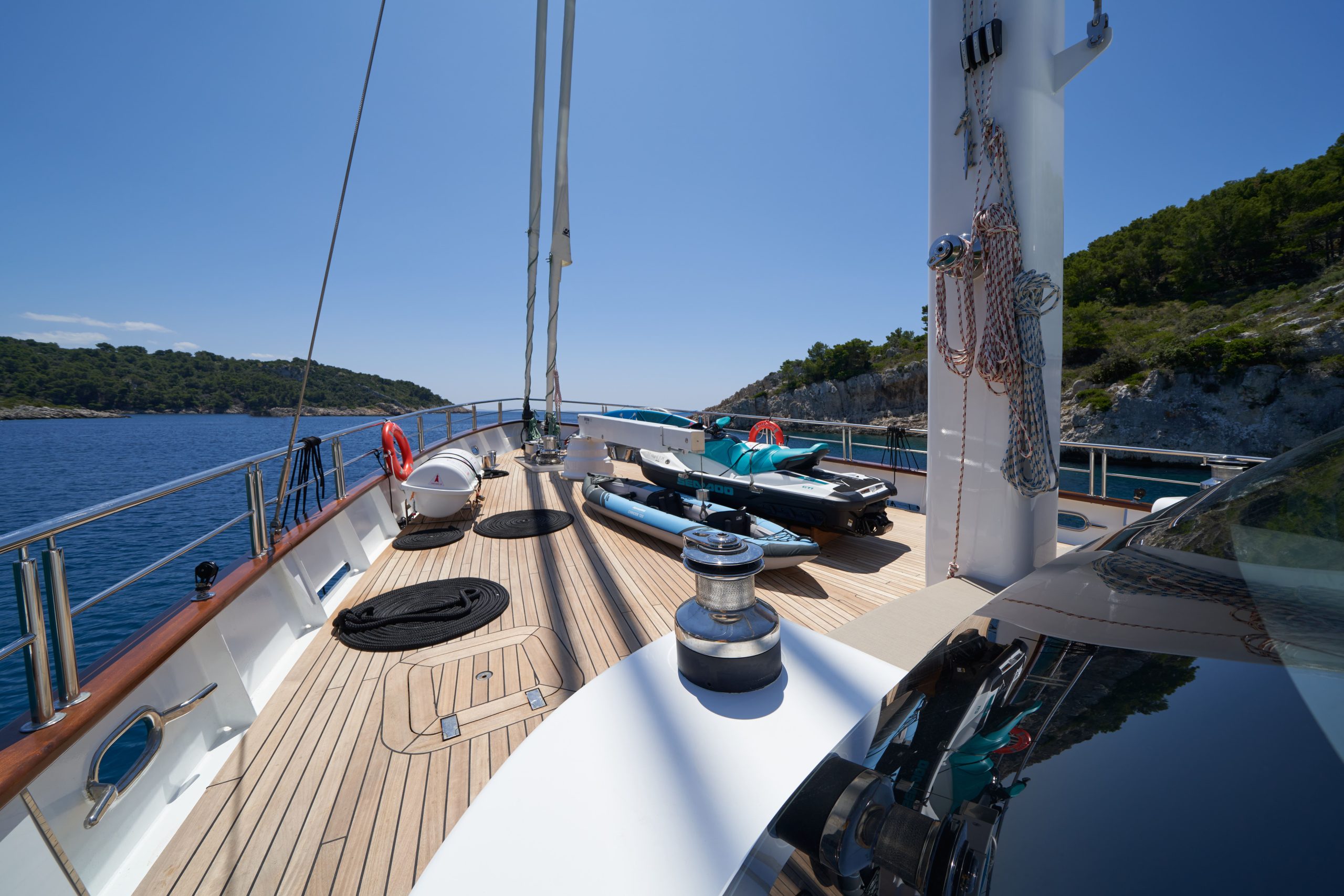 MARALLURE The image shows the deck of a yacht with polished wood flooring, coiled ropes, and lifebuoys. The yacht is docked near a rocky, green shoreline under a clear blue sky on a sunny day.